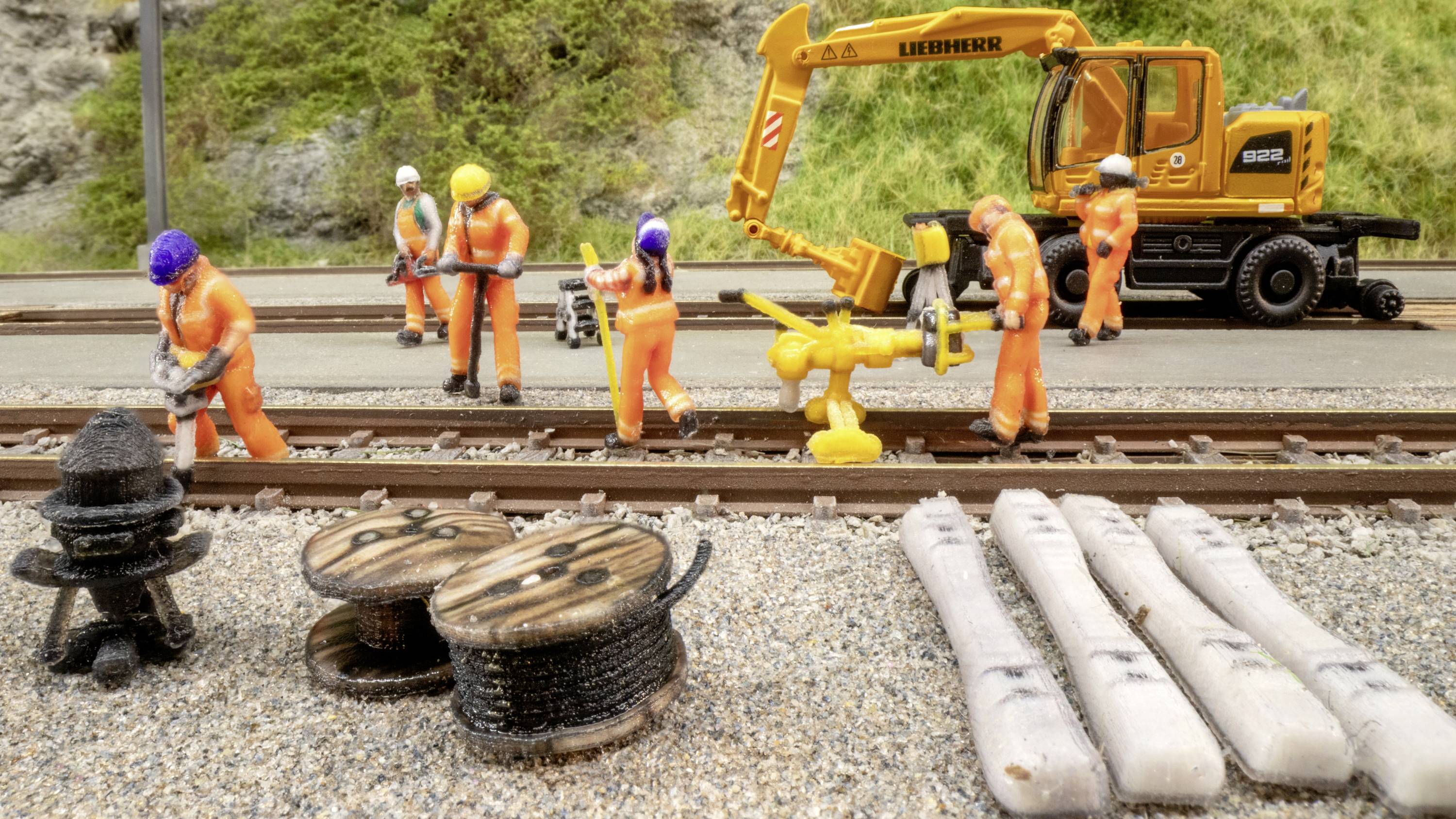 Des travailleurs en vêtements de protection orange réparent des voies ferrées. Un pelleteuse jaune se trouve en arrière-plan. Au premier plan, des bobines de câbles et des traverses.