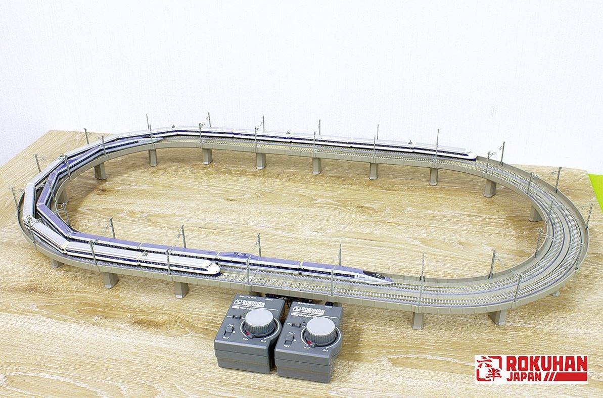 Ligne de chemin de fer miniature sur une table en bois, avec deux contrôleurs au premier plan, logo 'Rokuhan Japan' en bas à droite.