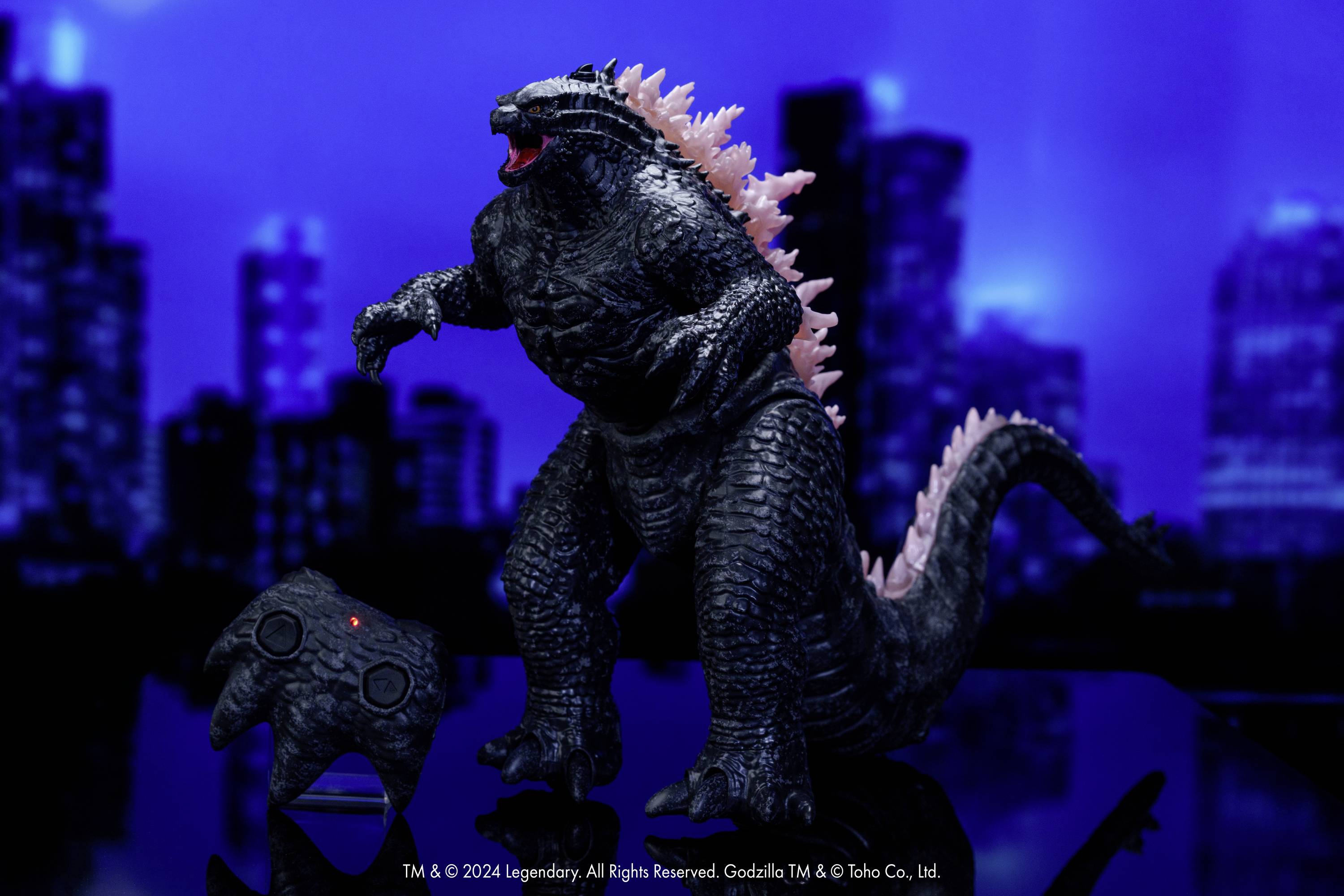 Une figurine de Godzilla noire avec des piquants roses se dresse devant un décor de gratte-ciels flous. Un petit contrôleur est posé en bas à gauche.