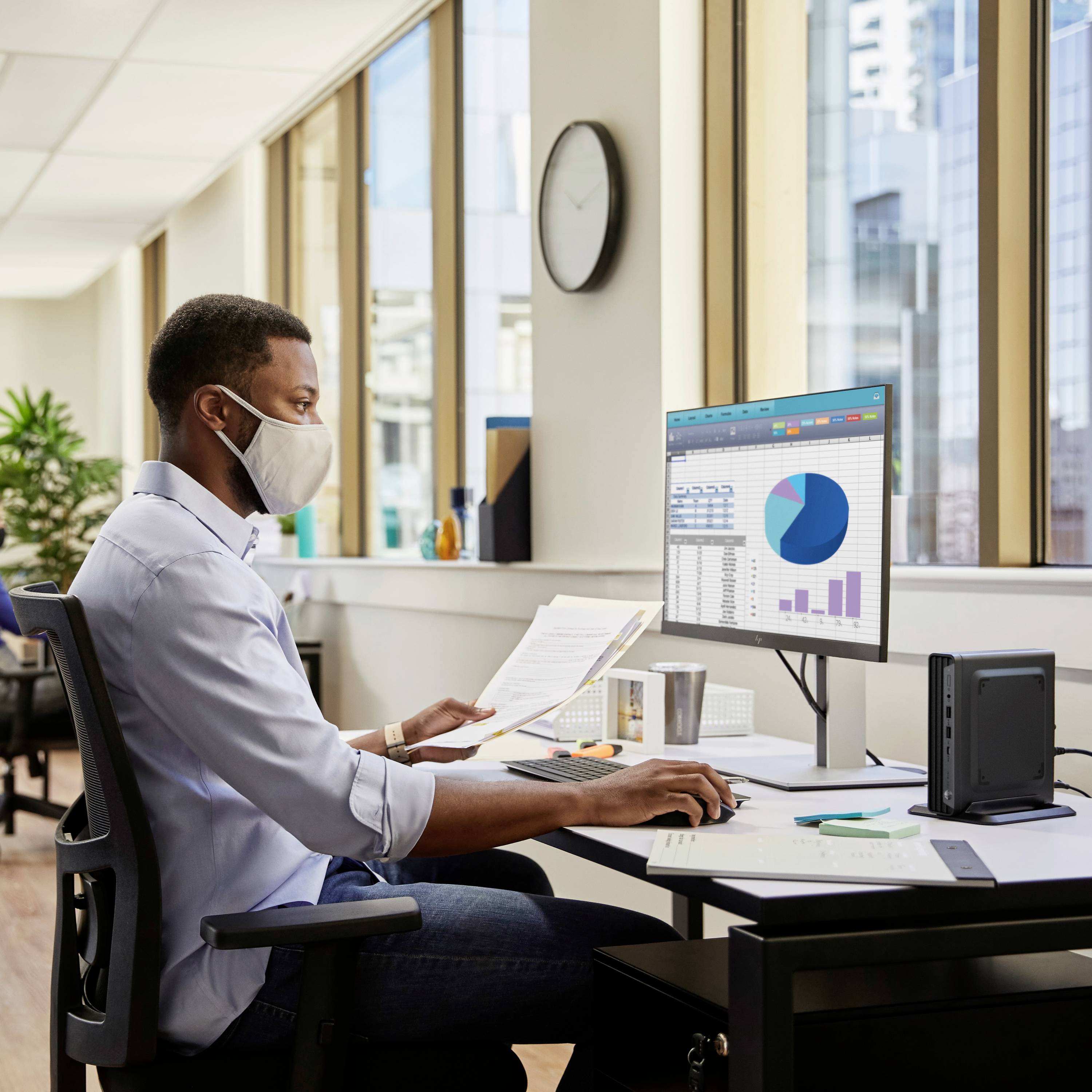 Un homme portant un masque est assis à un bureau avec un ordinateur. Sur l'écran, on peut voir des diagrammes et des tableaux colorés.
