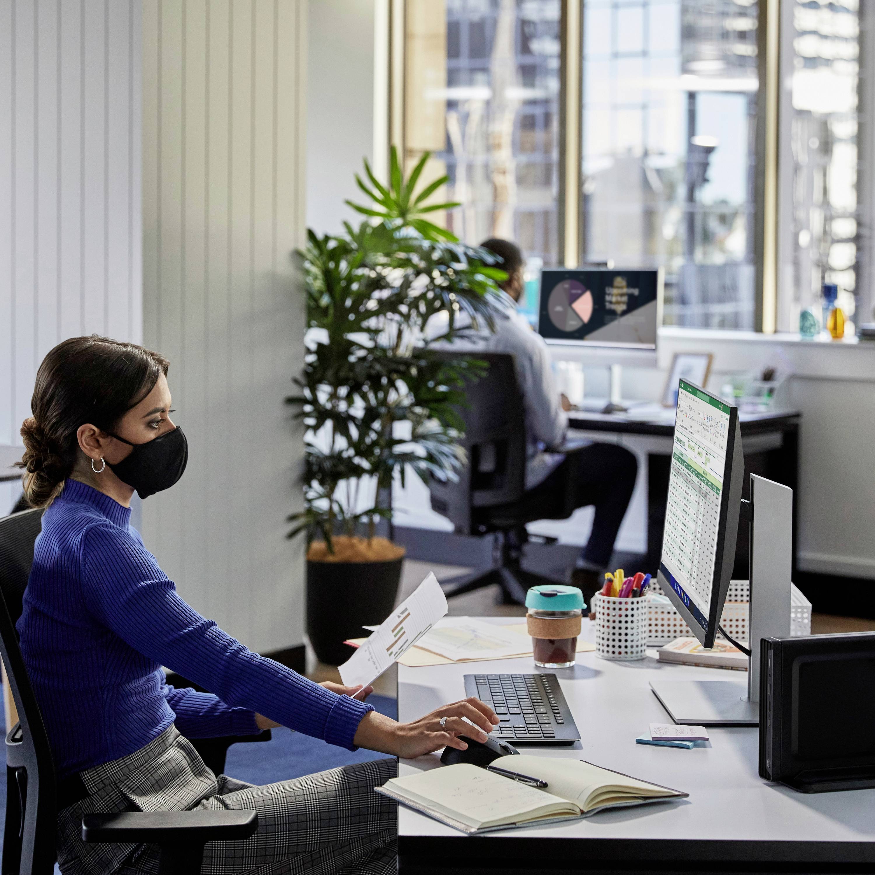 Une femme portant un masque travaille à un bureau avec un ordinateur, un papier à la main. À l'arrière-plan, un homme est assis à un second bureau.