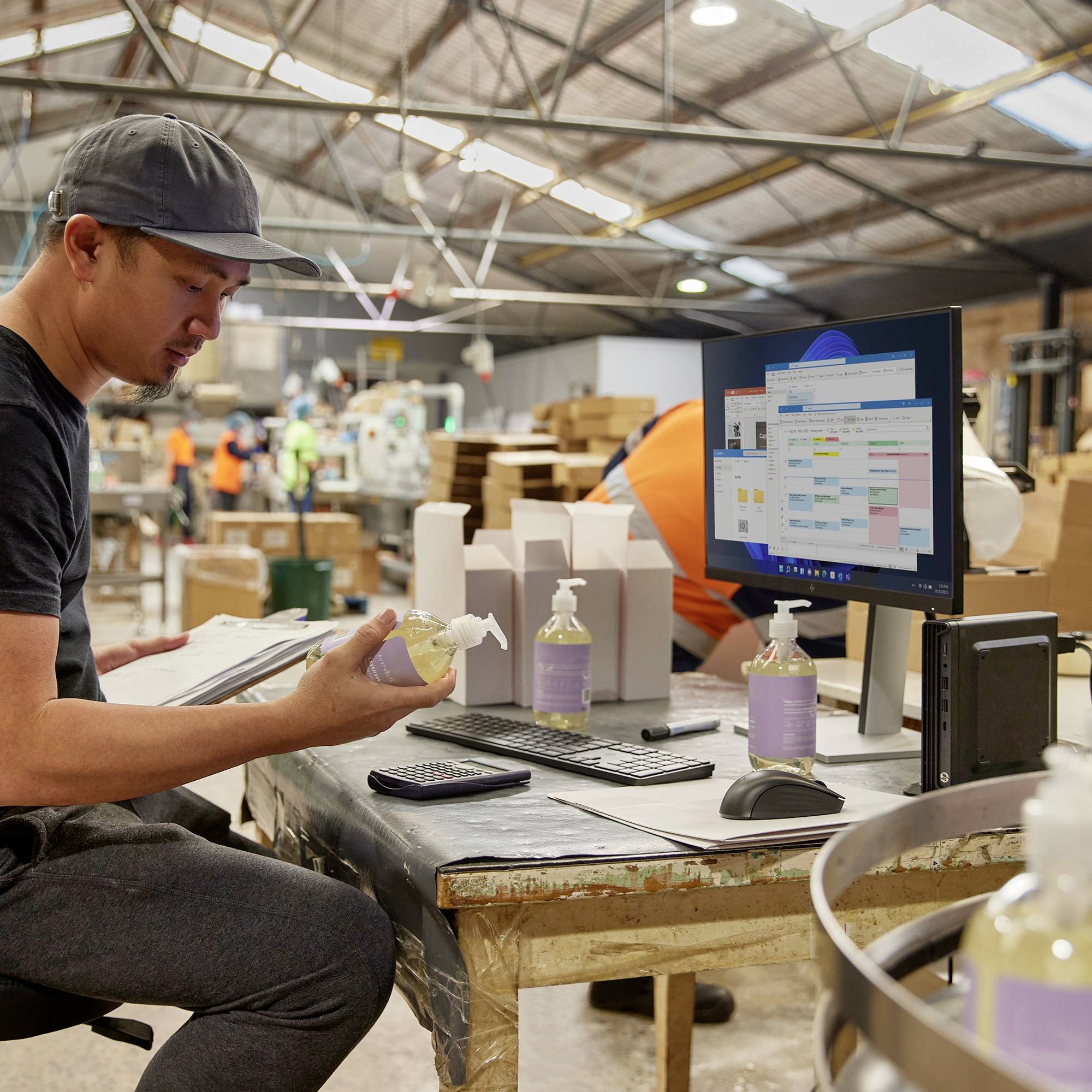 Un homme portant une casquette est assis dans une halle d'usine à une table sur laquelle se trouvent un écran d'ordinateur avec un plan et du gel hydroalcoolique. À l'arrière-plan, d'autres employés travaillent sur des machines.