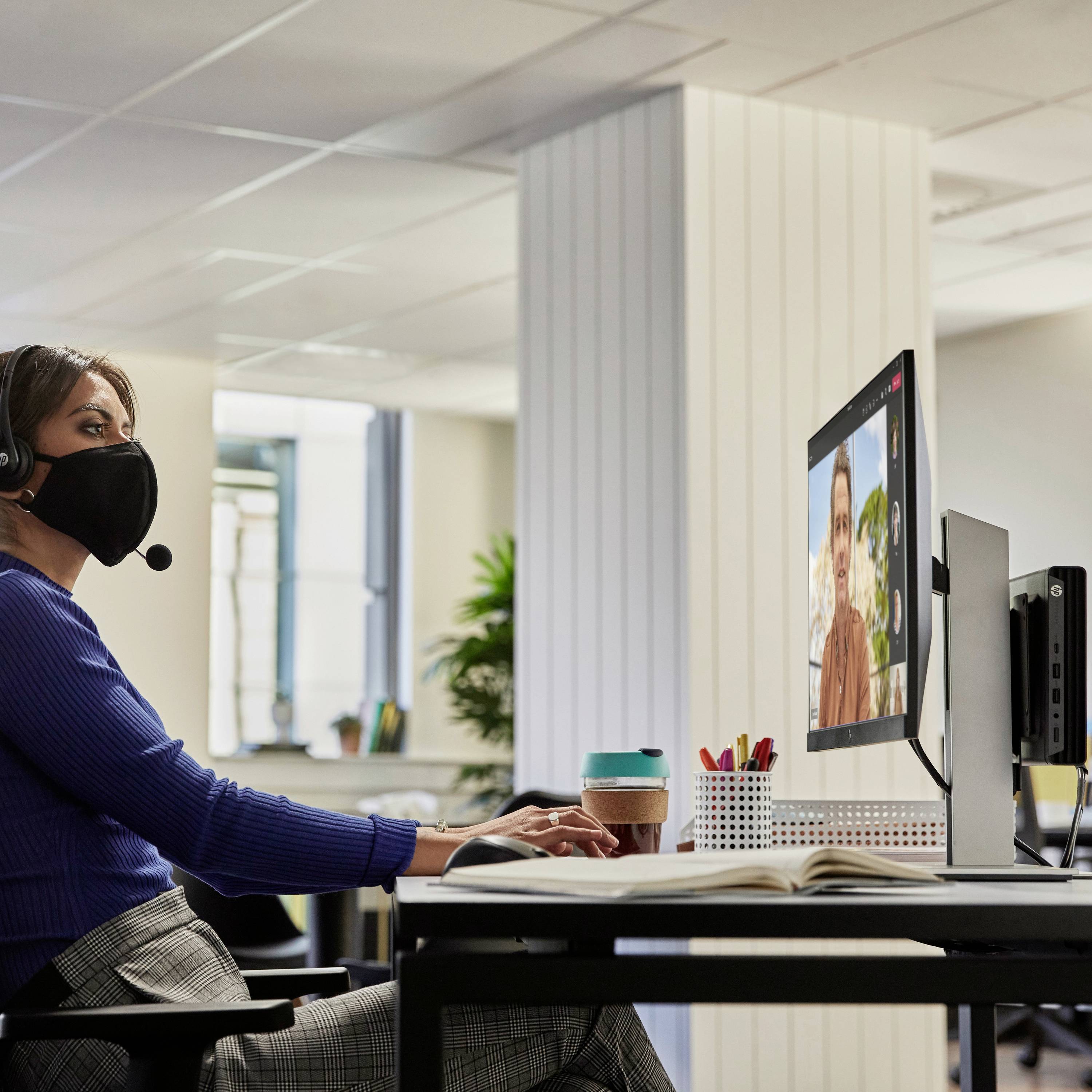 Une personne portant un masque est assise à un bureau et participe à un appel vidéo sur un écran d'ordinateur.