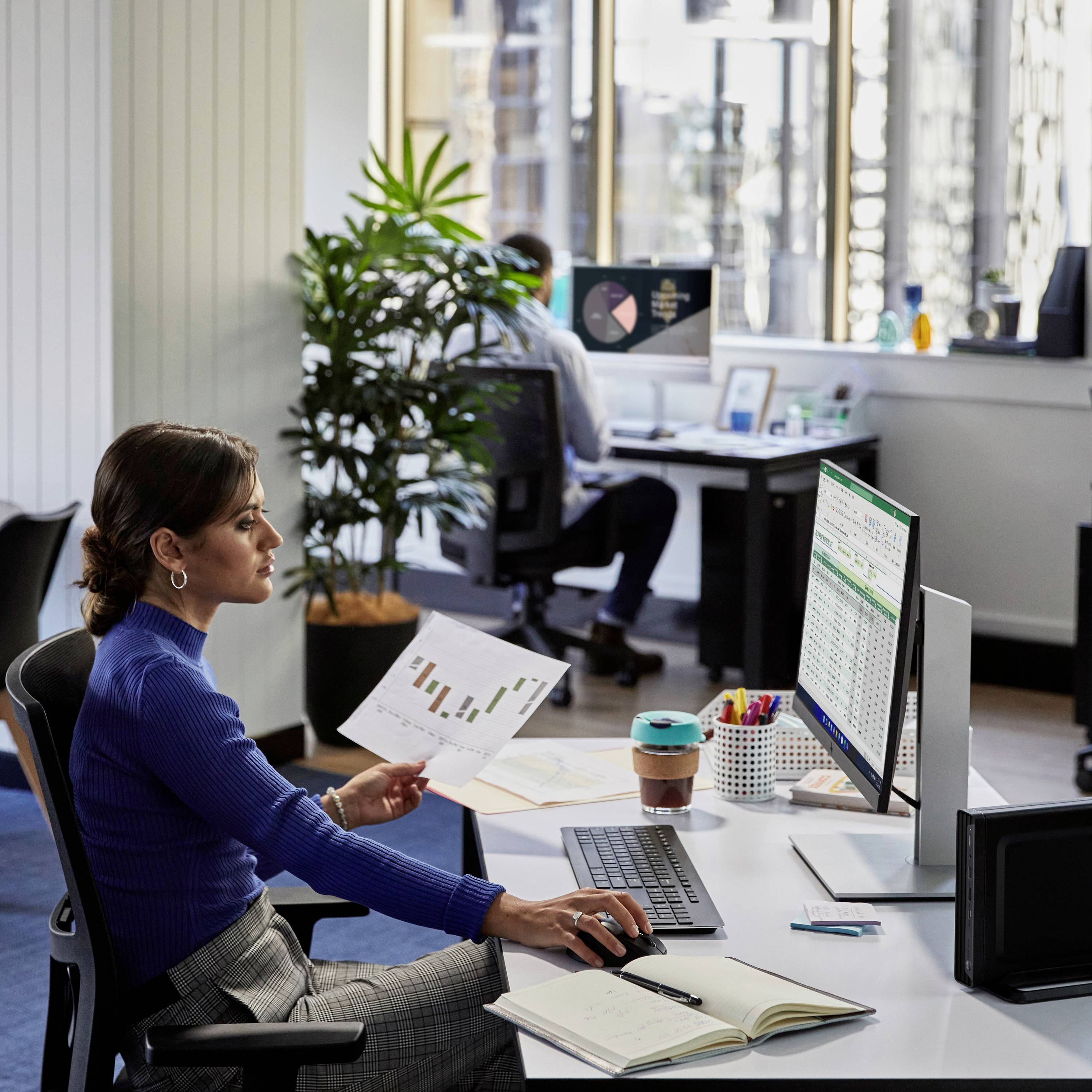 Une femme dans un bureau examine une feuille avec des diagrammes. Un moniteur affichant des tableurs est posé devant elle sur le bureau. À l'arrière-plan, une personne travaille sur un ordinateur.