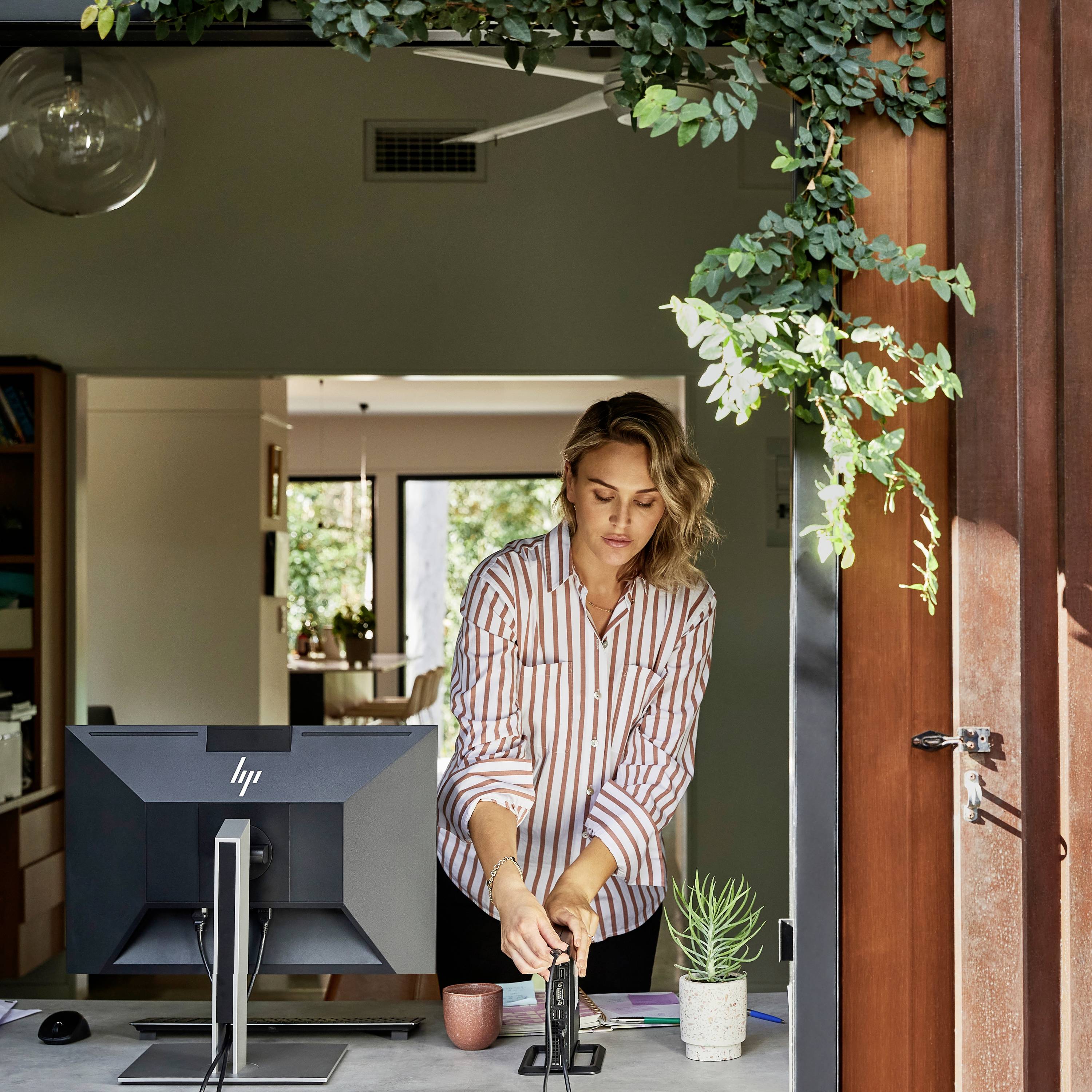 Une femme est debout à un bureau avec un ordinateur et un smartphone, des plantes suspendues descendent du plafond.