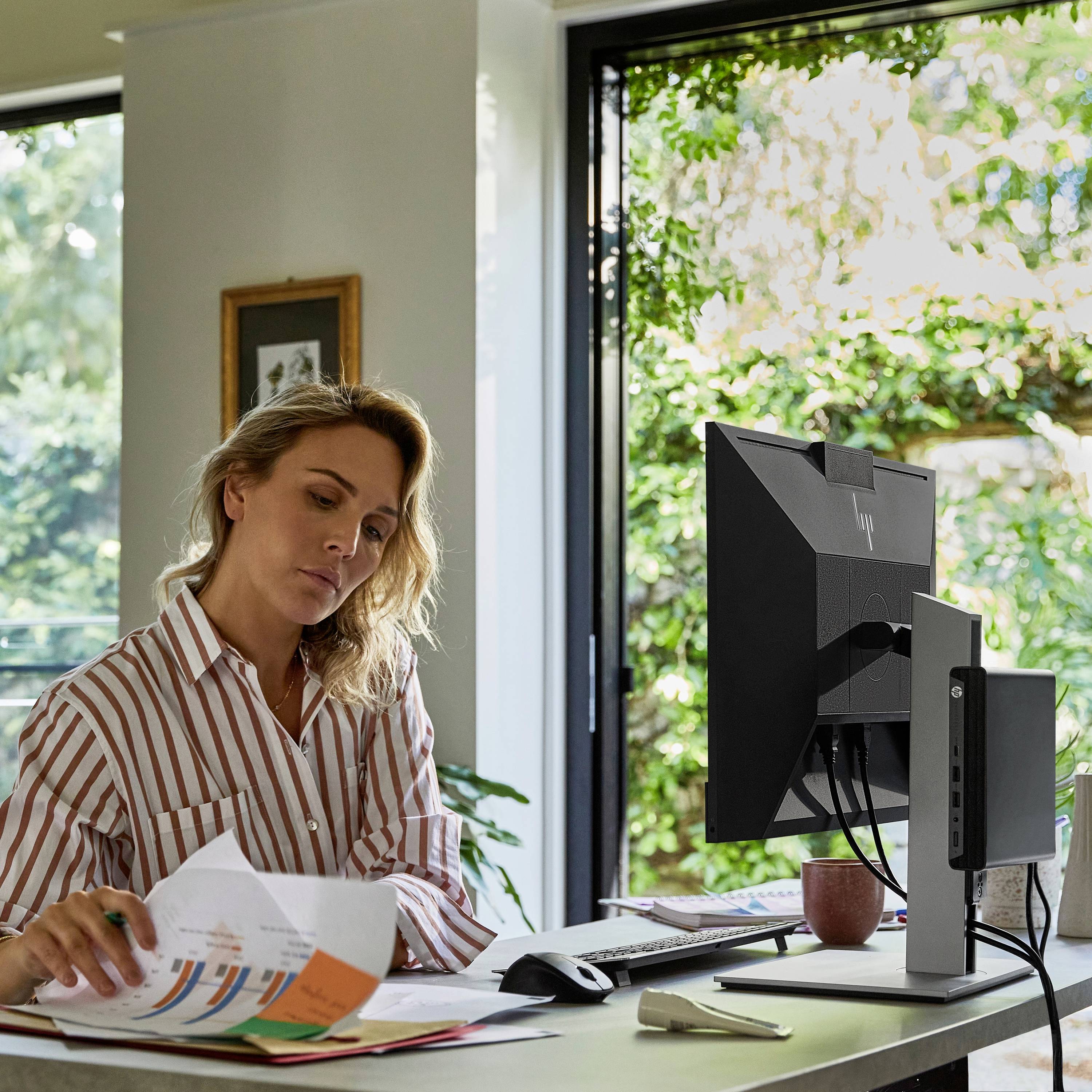 Une personne est assise à un bureau, examine des documents et utilise un ordinateur. La pièce est éclairée par une lumière naturelle.