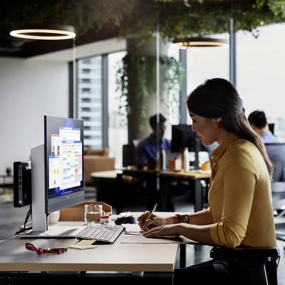 Une personne portant une chemise jaune travaille sur un ordinateur dans un bureau moderne avec plusieurs personnes en arrière-plan.