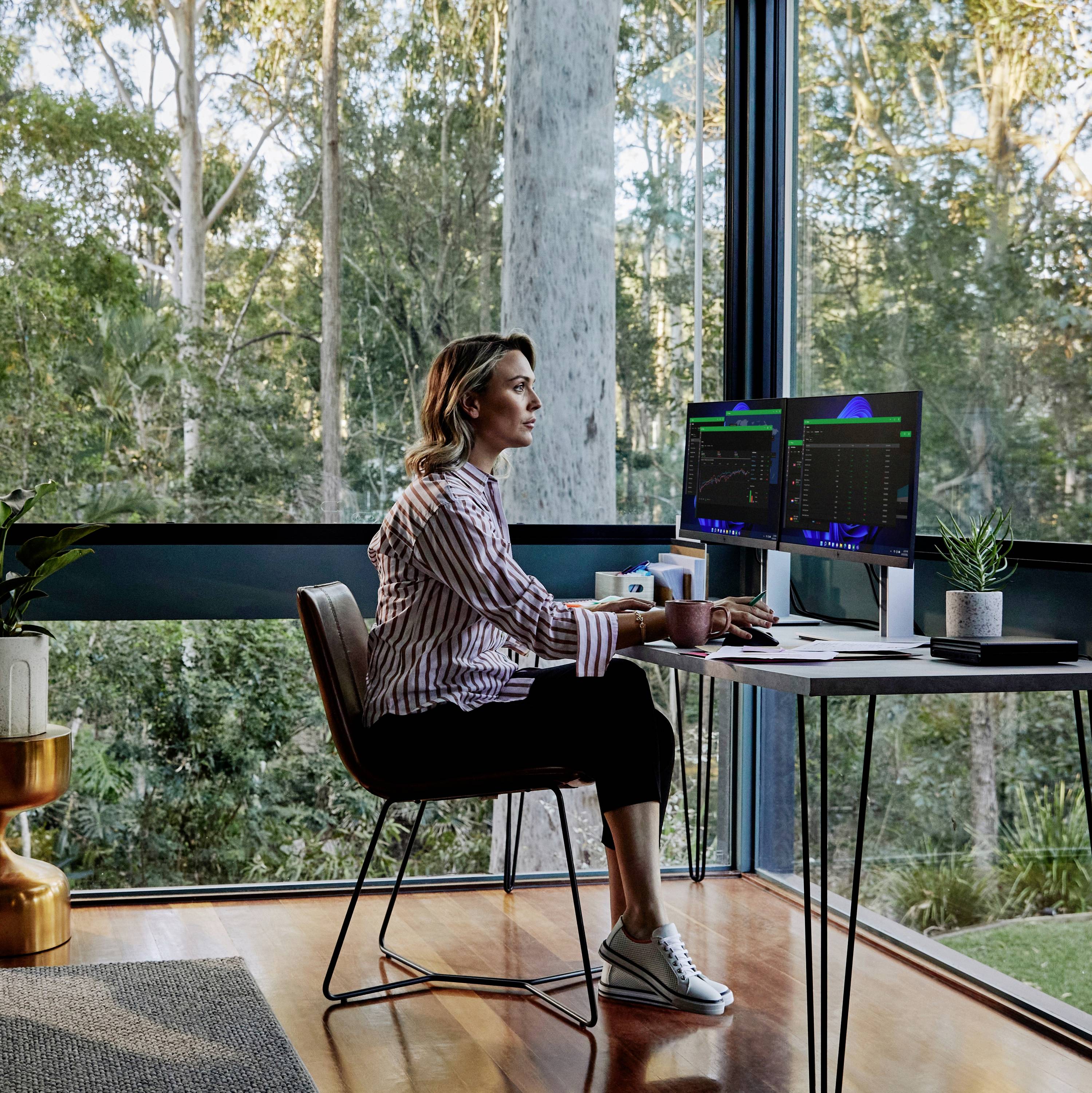 Une personne travaille à un bureau avec plusieurs écrans et un ordinateur portable, entourée de grandes fenêtres donnant sur une forêt.