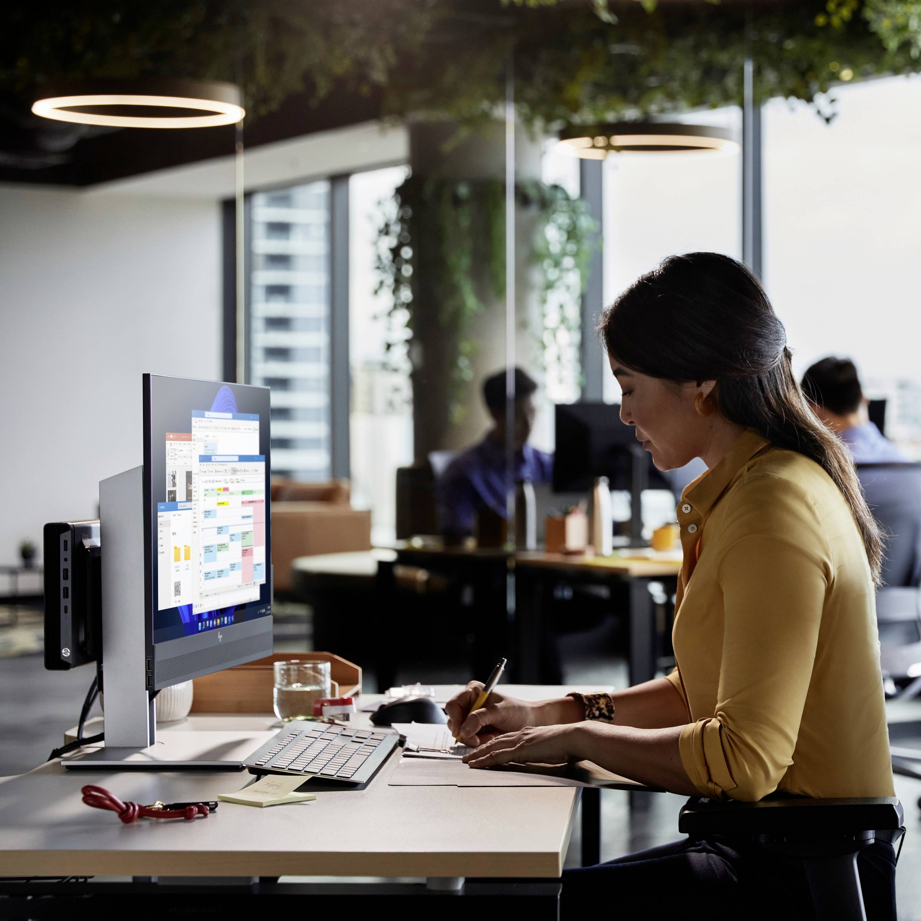 Une femme est assise à un bureau dans un bureau moderne, travaillant sur un ordinateur, tandis que des personnes travaillent en arrière-plan.