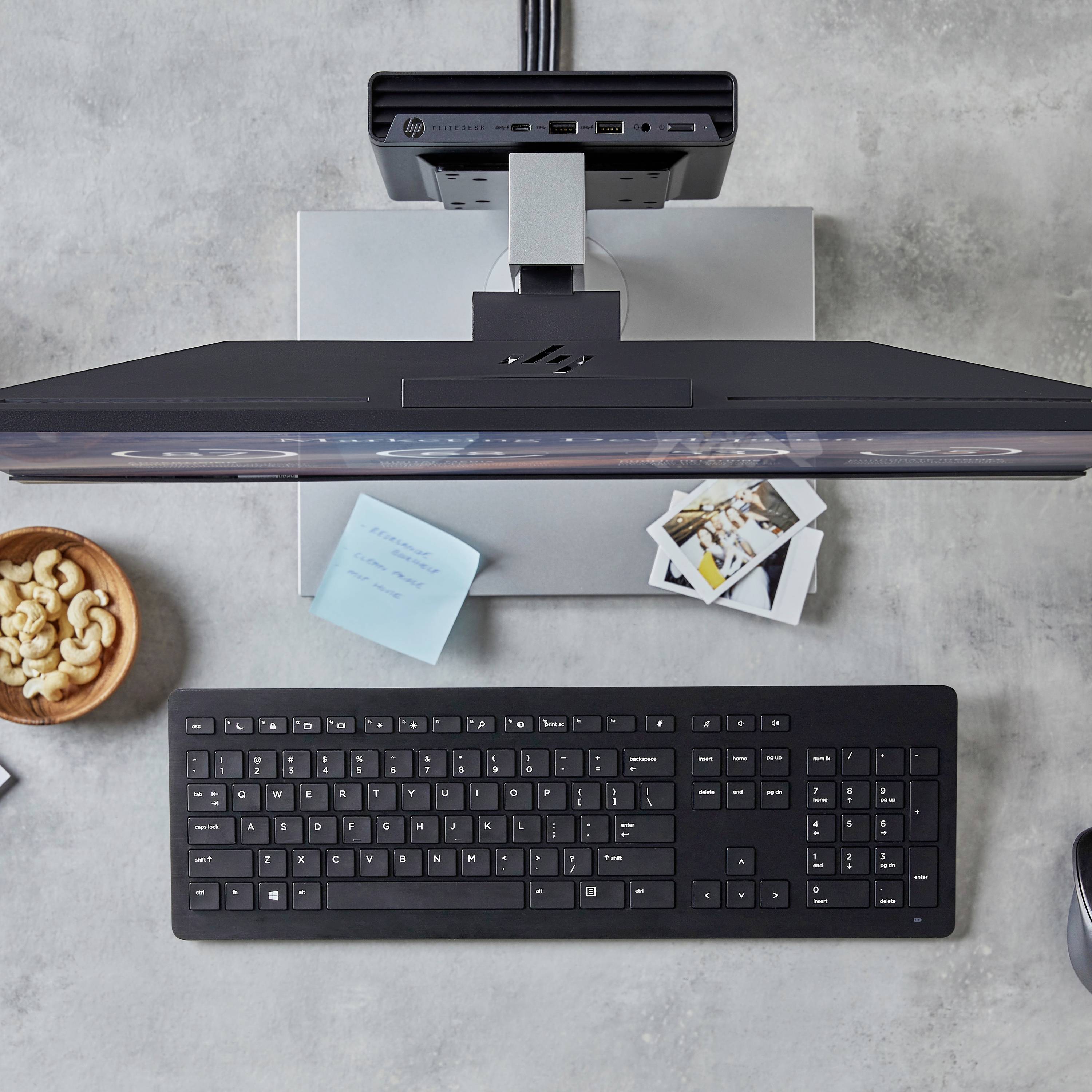 Un bureau avec un clavier d'ordinateur, un écran, un bloc-notes, des photos et une assiette de noix de cajou.