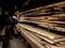 Un homme mesure de grandes planches de bois avec un mètre ruban dans un entrepôt. À l'arrière-plan, des planches de bois sont empilées.