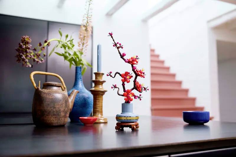 Une nature morte soigneusement composée avec une théière décorative, des bougeoirs et une branche fleurie dans un vase posés sur une table.