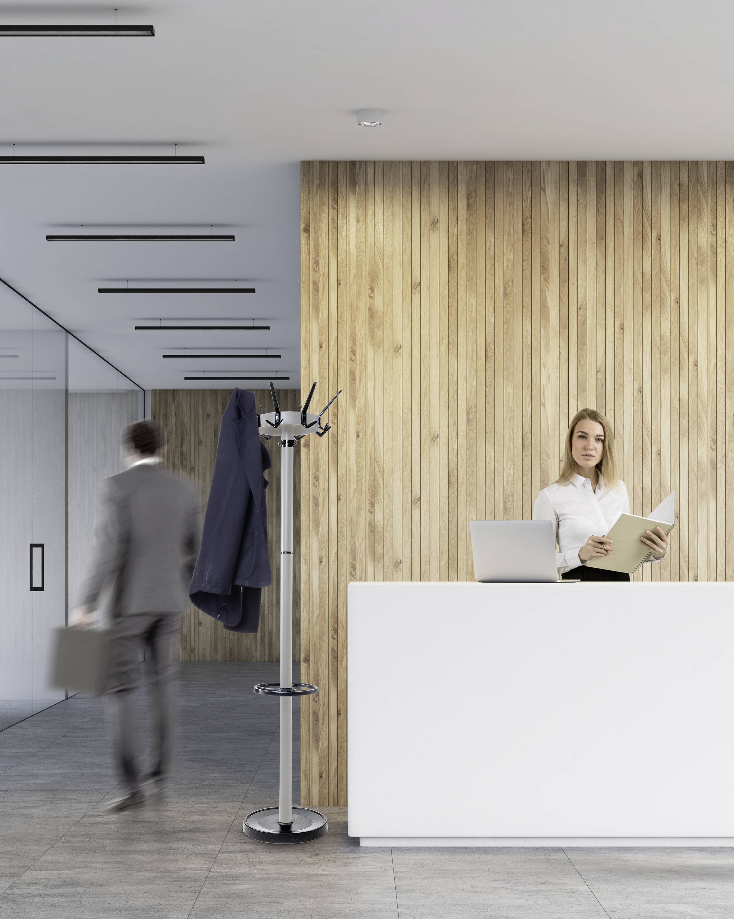 Une femme se tient derrière un comptoir de réception dans un bureau moderne. Un homme en costume passe à côté. Une paroi en bois est visible en arrière-plan.