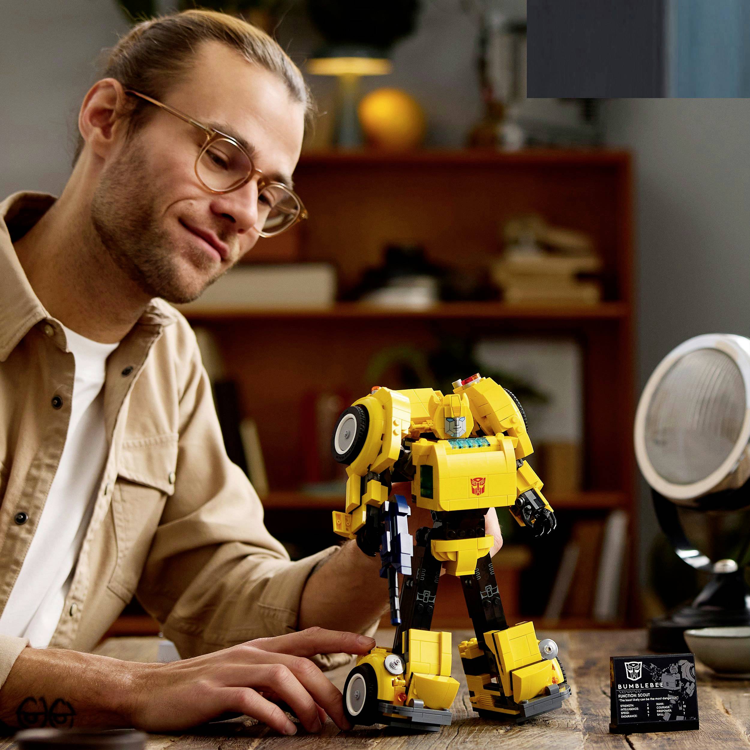 Une personne regarde et touche une voiture jouet jaune en forme de robot qui est posée sur une table. En arrière-plan, on aperçoit des étagères.