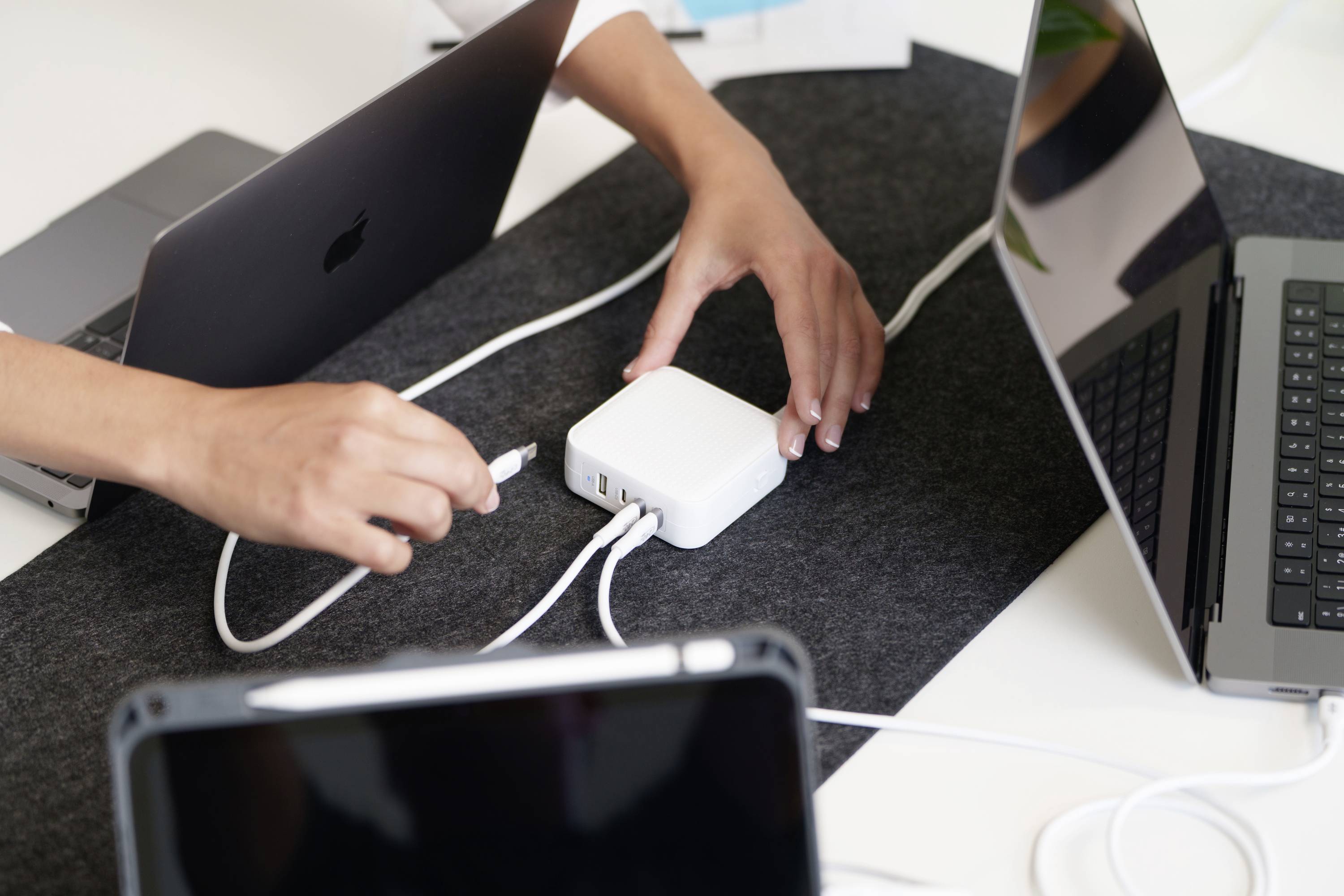 Une personne branche un câble de charge dans un hub USB blanc posé sur une table à côté de deux ordinateurs portables et d'une tablette.