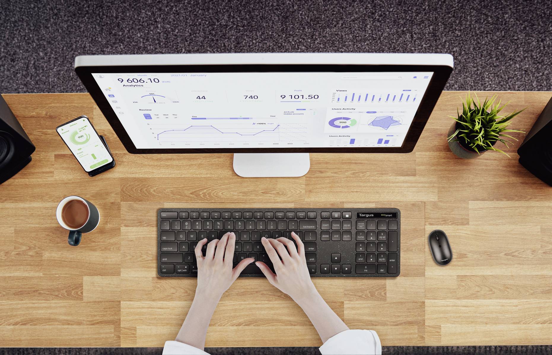Vue d'ensemble : Une personne tapant sur un clavier devant un écran affichant des diagrammes et des statistiques ; un smartphone, une tasse de café et une plante à proximité.