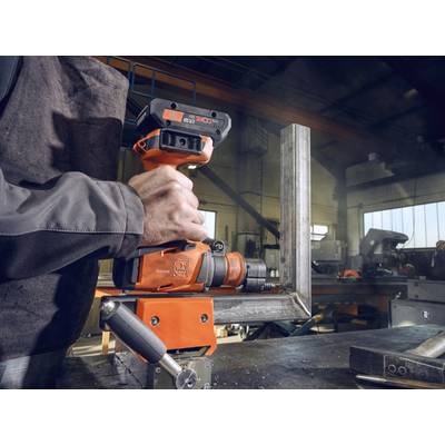 Une personne tient une perceuse orange dans un atelier. Des cadres métalliques et des machines sont visibles en arrière-plan.