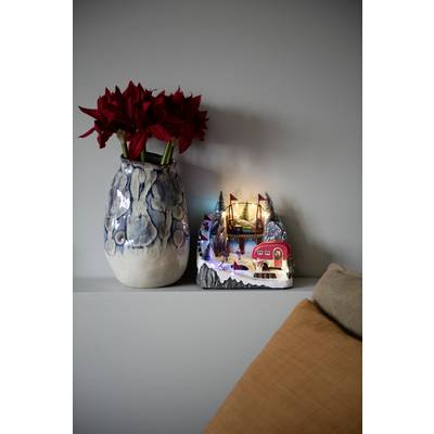 'Vase avec des fleurs rouges et village miniature éclairé sur une étagère, à côté d'un coussin beige. Ambiance chaleureuse et accueillante.'