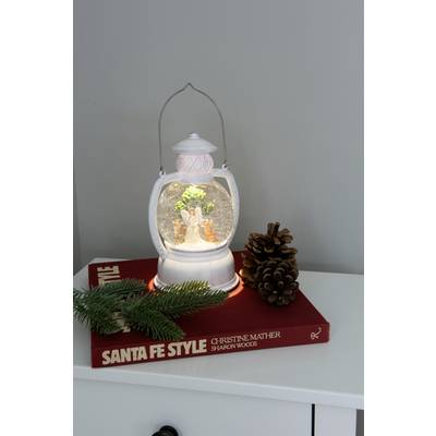 Une lanterne blanche avec un motif de boule à neige, posée sur un livre rouge portant le titre 'Santa Fe Style'. À côté, deux pommes de pin et une branche de sapin.