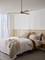 Une chambre au design moderne avec un lit décoré de plusieurs coussins et d'une couverture beige. Des murs clairs et un ventilateur de plafond complètent le design minimaliste.