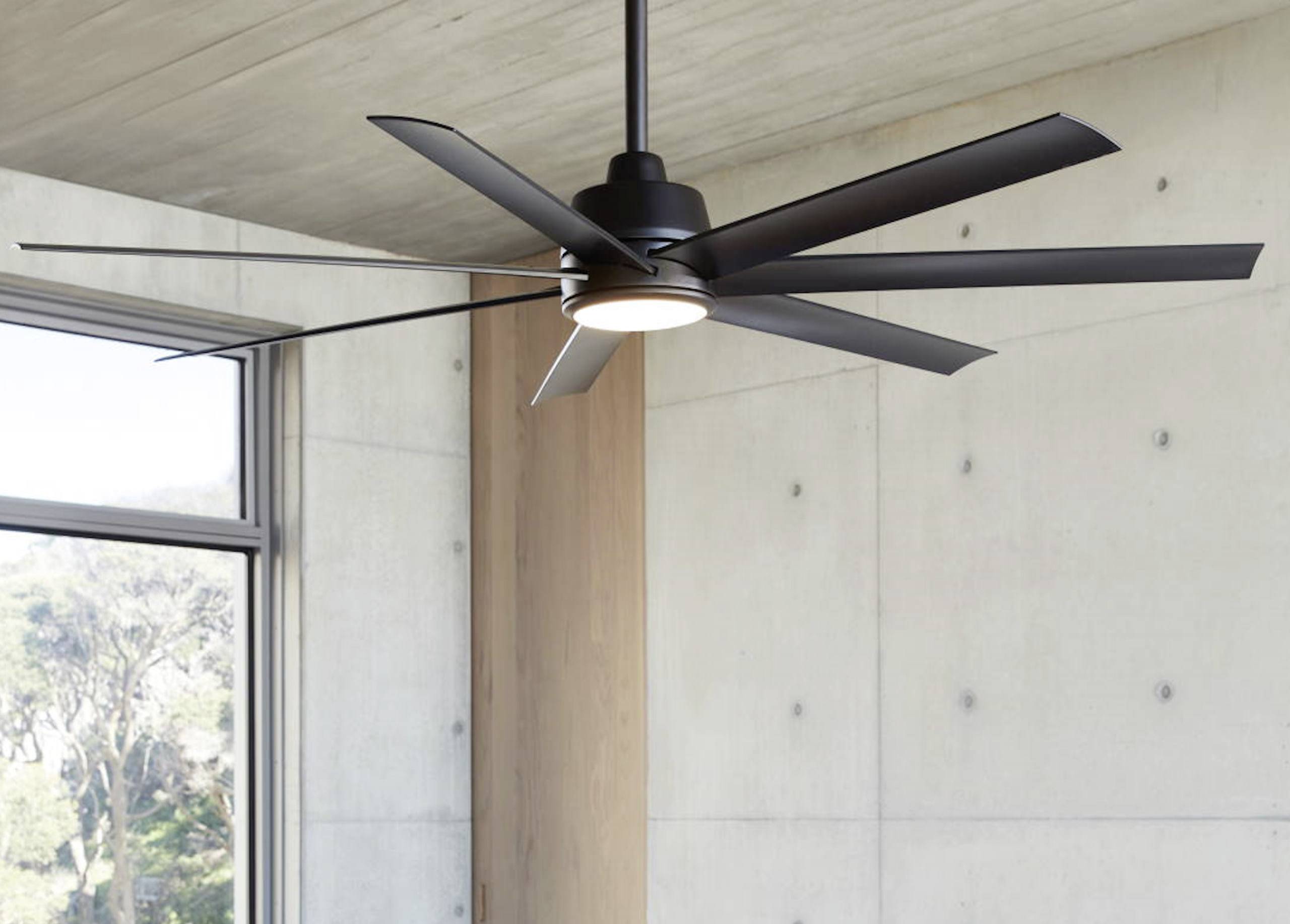 Un ventilateur de plafond moderne à plusieurs pales est suspendu au plafond dans une pièce lumineuse avec de grandes fenêtres.