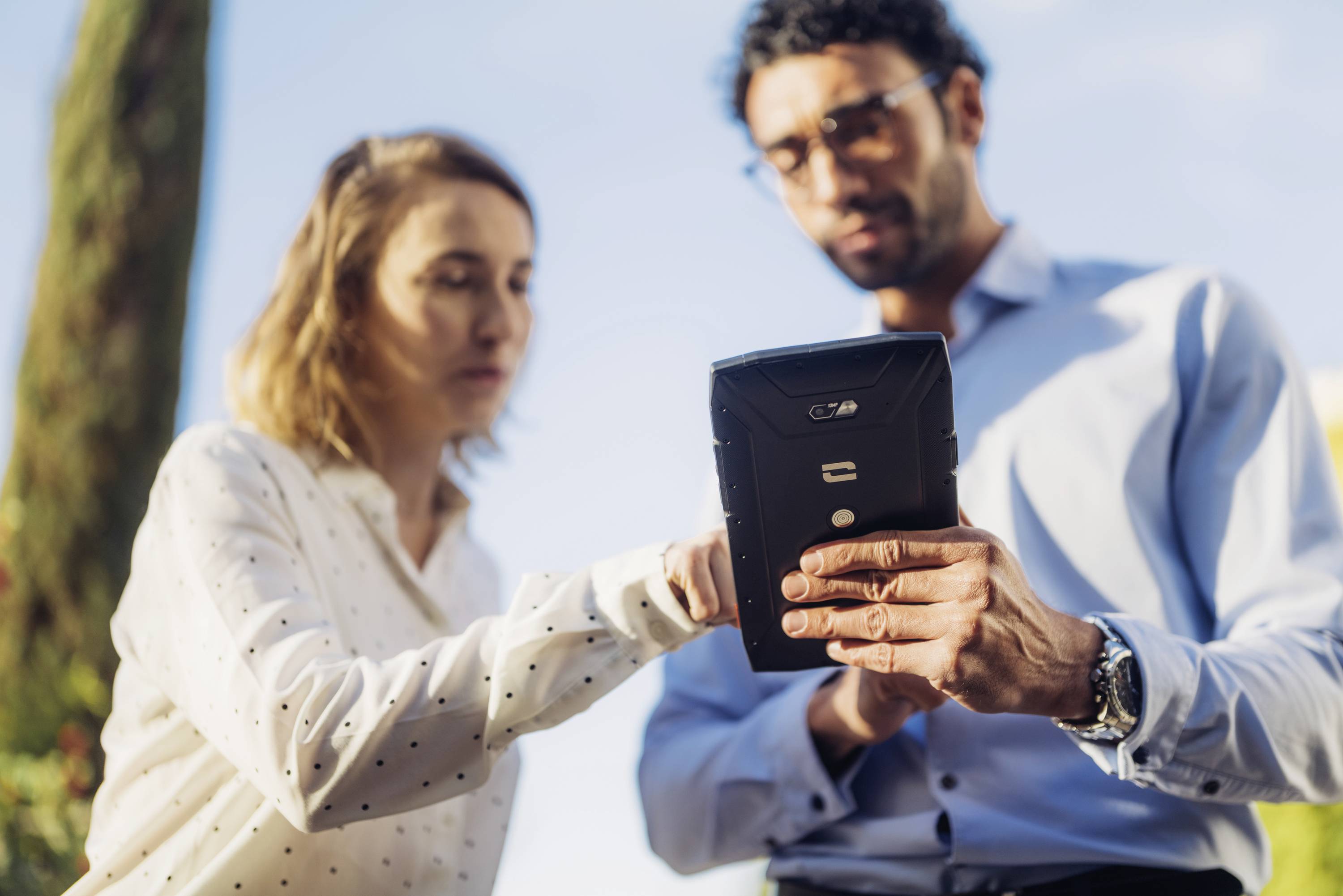 Deux personnes sont dehors et regardent une tablette. L'une d'entre elles pointe l'écran du doigt. Elles semblent concentrées et intéressées.