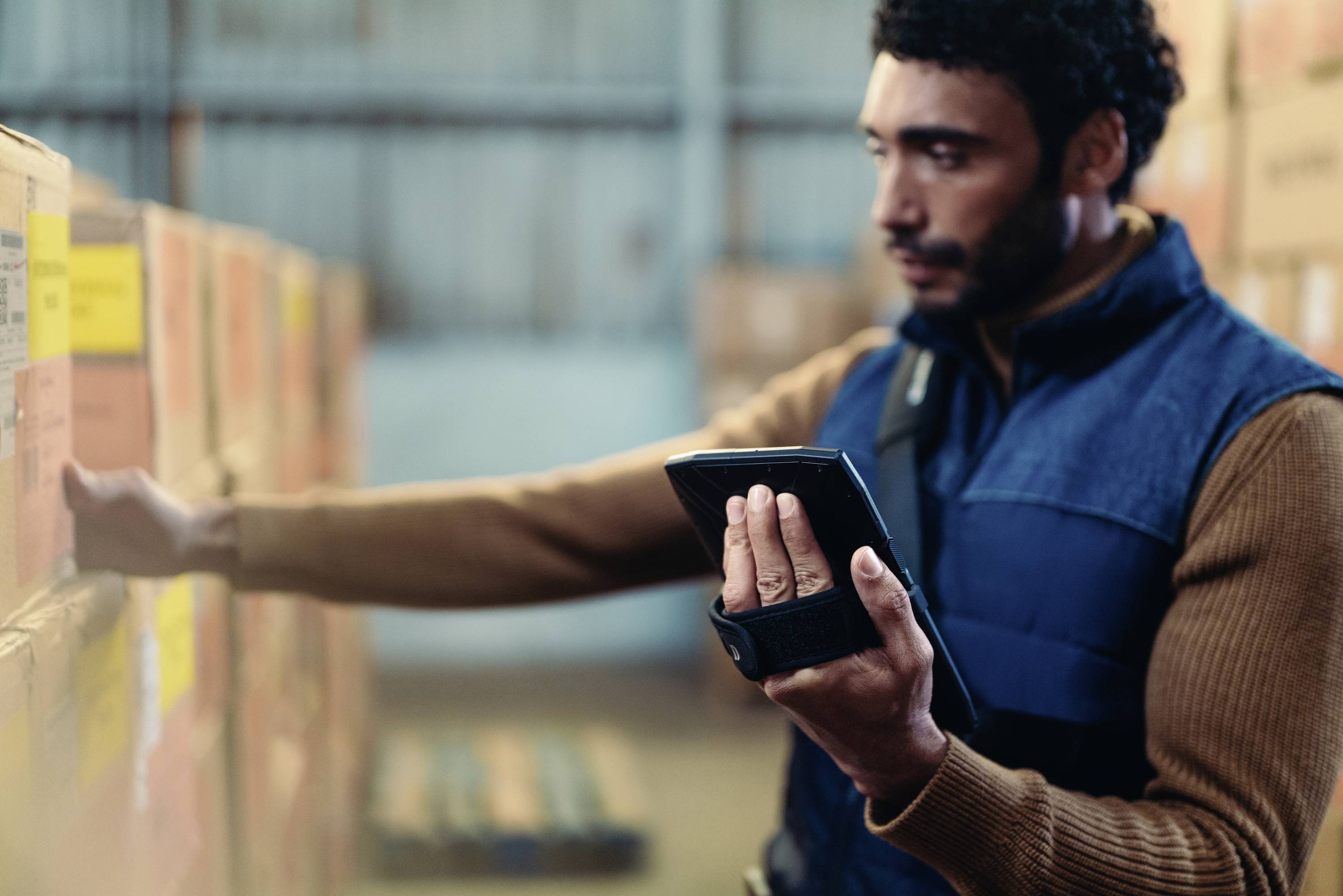 Un homme vérifie les stocks dans un entrepôt à l'aide d'une tablette. Il se trouve devant des étagères remplies de cartons.