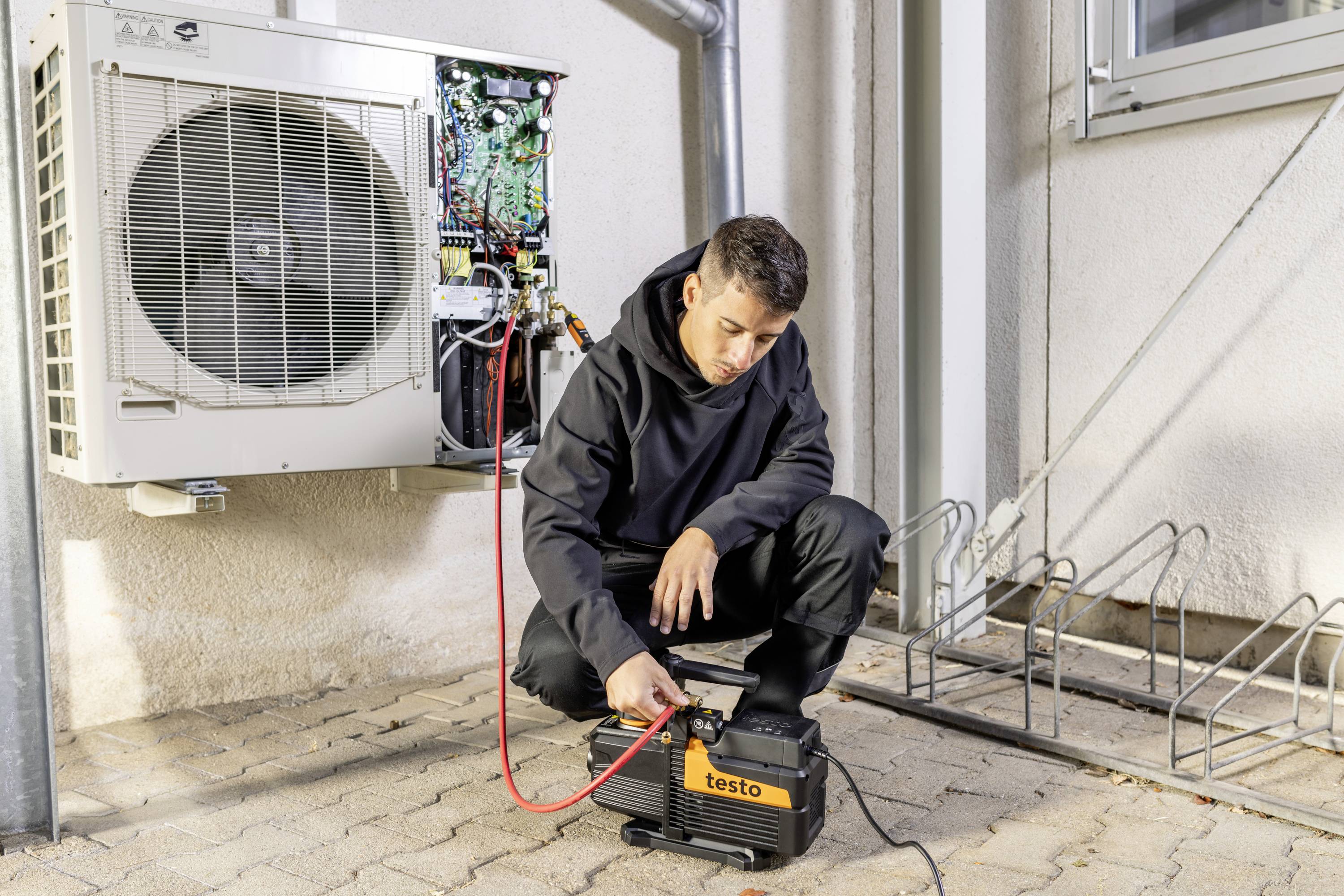 Un homme vérifie un climatiseur à l'extérieur. Il utilise un appareil de test et est à genoux devant l'appareil sur un sol pavé.