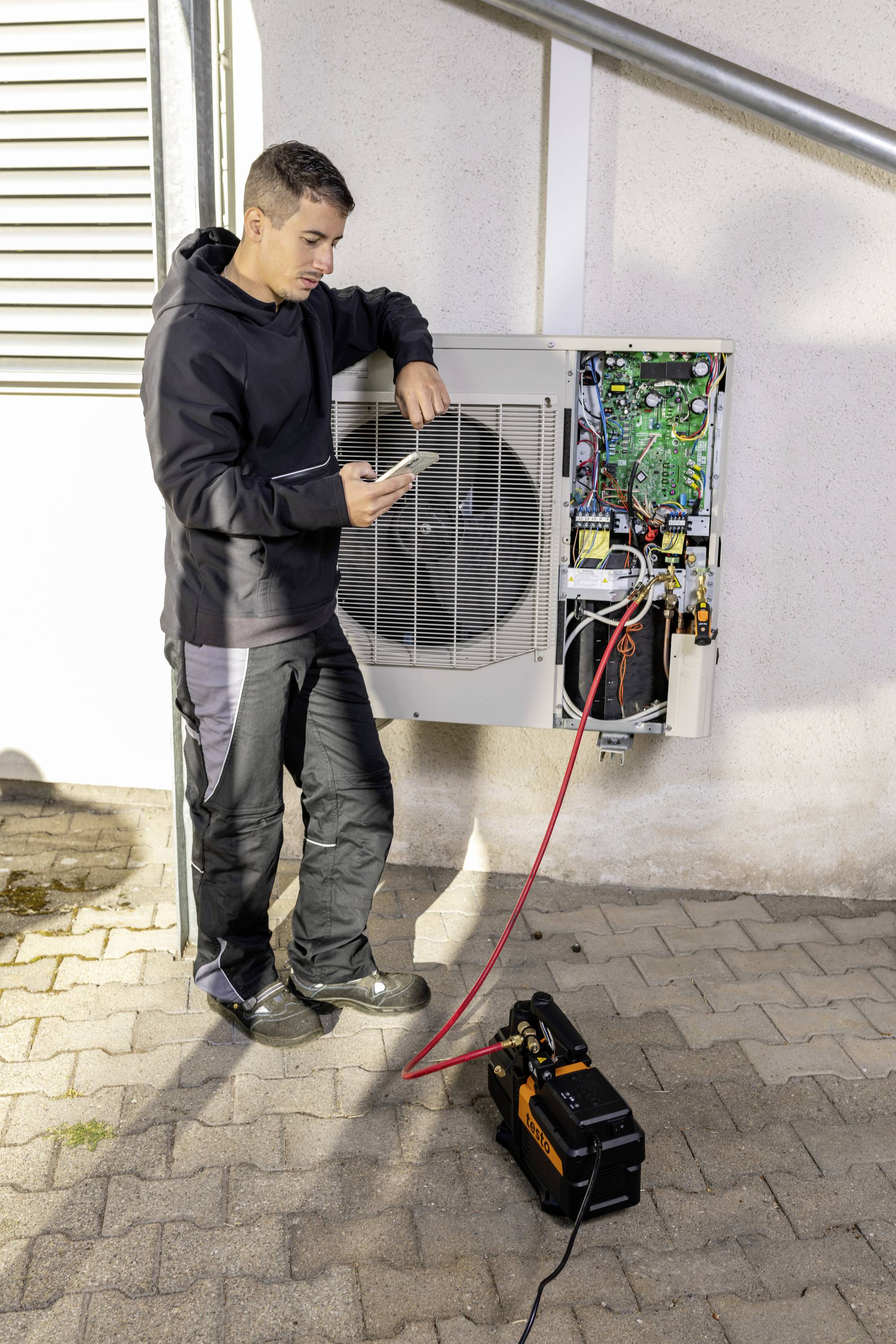 Un technicien vérifie un appareil extérieur de climatisation ouvert à l'aide d'une tablette. Un dispositif de test y est connecté.