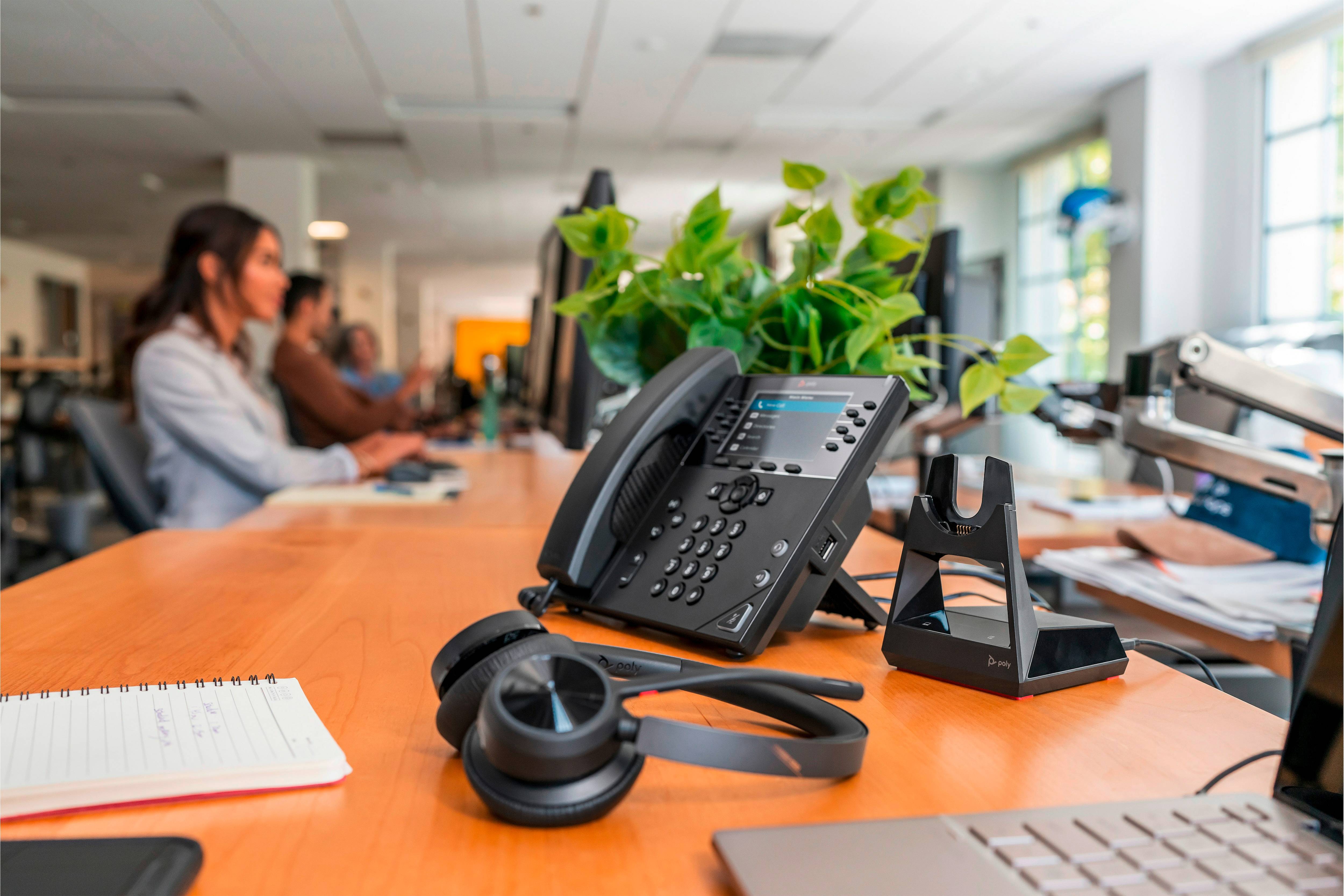 Un téléphone et un casque sont posés sur un bureau dans un bureau animé. En arrière-plan, des personnes travaillent sur des ordinateurs.