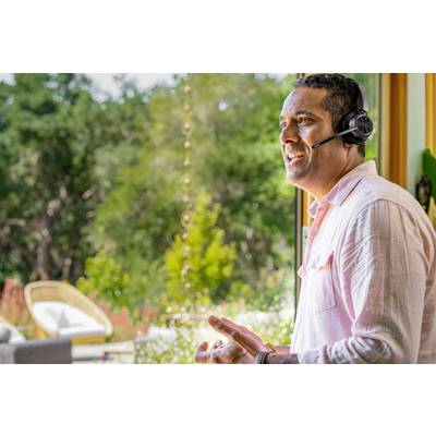 Un homme portant un casque téléphone en souriant dans une pièce lumineuse avec de grandes fenêtres. À l'extérieur, on peut voir des arbres et un jardin.