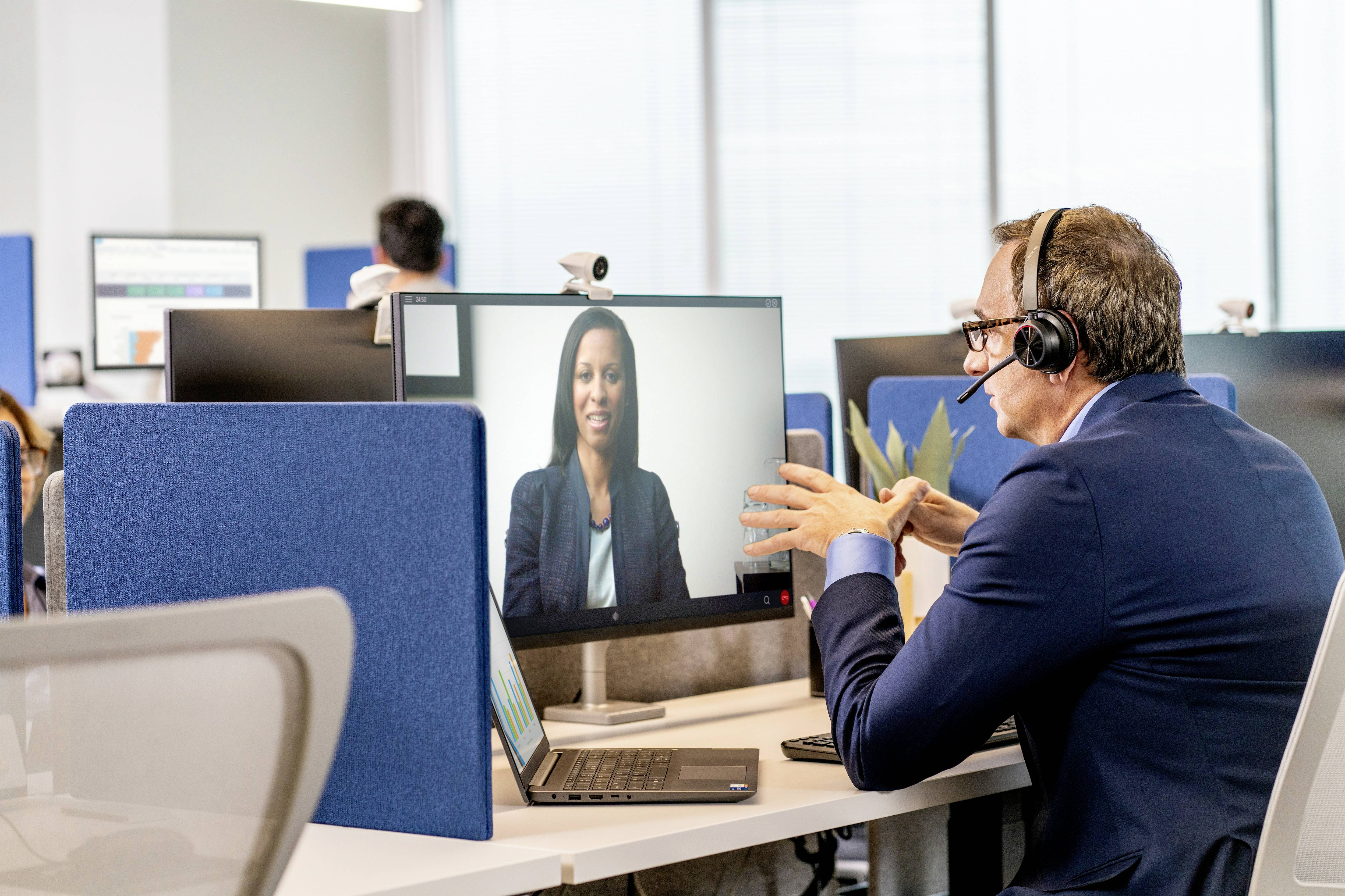 Un homme d'affaires portant un casque mène un appel vidéo sur ordinateur dans un bureau moderne. À l'arrière-plan, d'autres personnes travaillent.