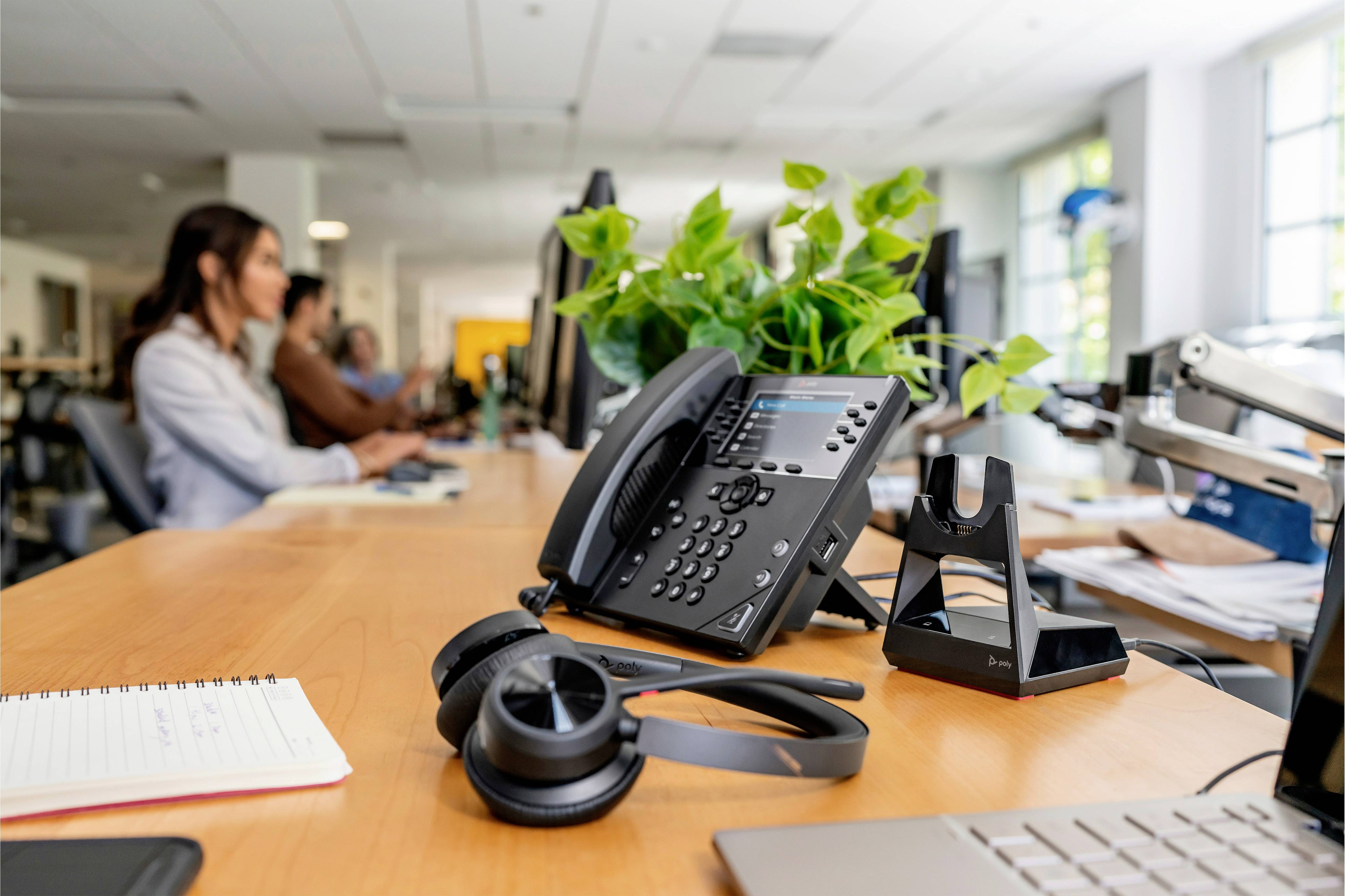 Un bureau dans un espace de travail avec un téléphone, des écouteurs, un bloc-notes et des plantes au premier plan. Des personnes travaillent en arrière-plan.