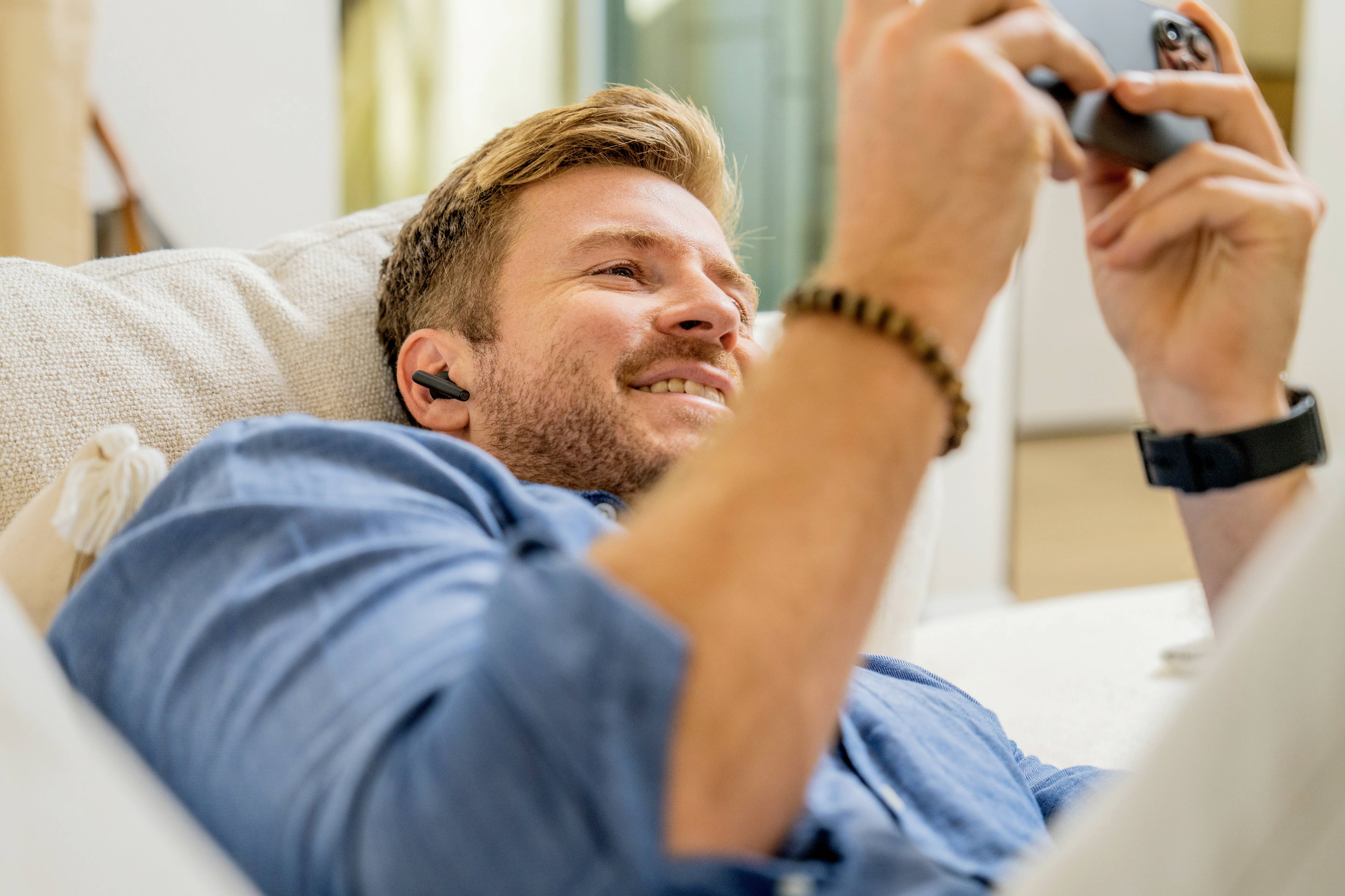 Un homme est allongé de manière détendue sur un canapé, porte des écouteurs sans fil et utilise un smartphone.
