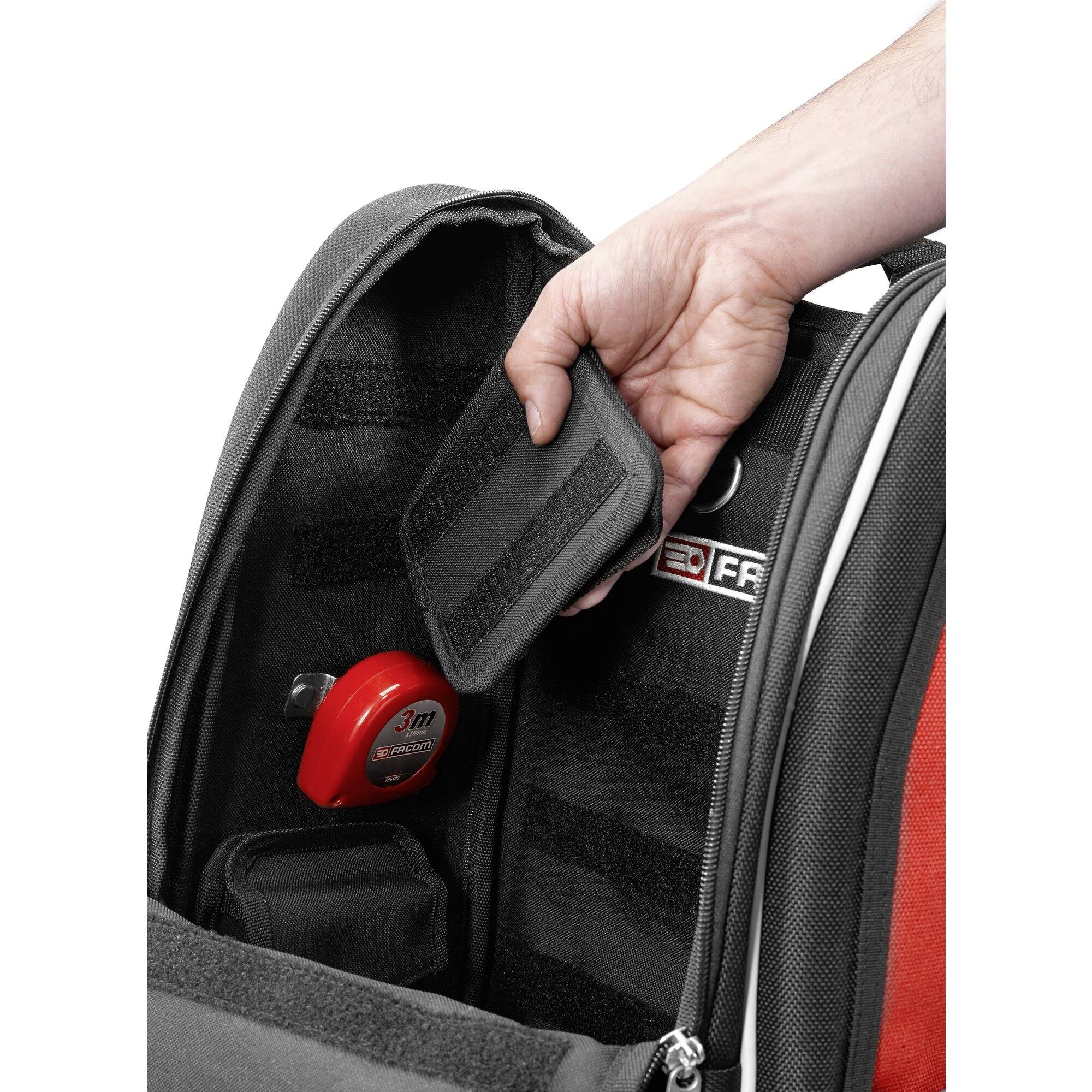 Une main ouvre une poche dans un sac à dos, où se trouve un mètre ruban rouge. Le sac à dos a des compartiments intérieurs noirs.