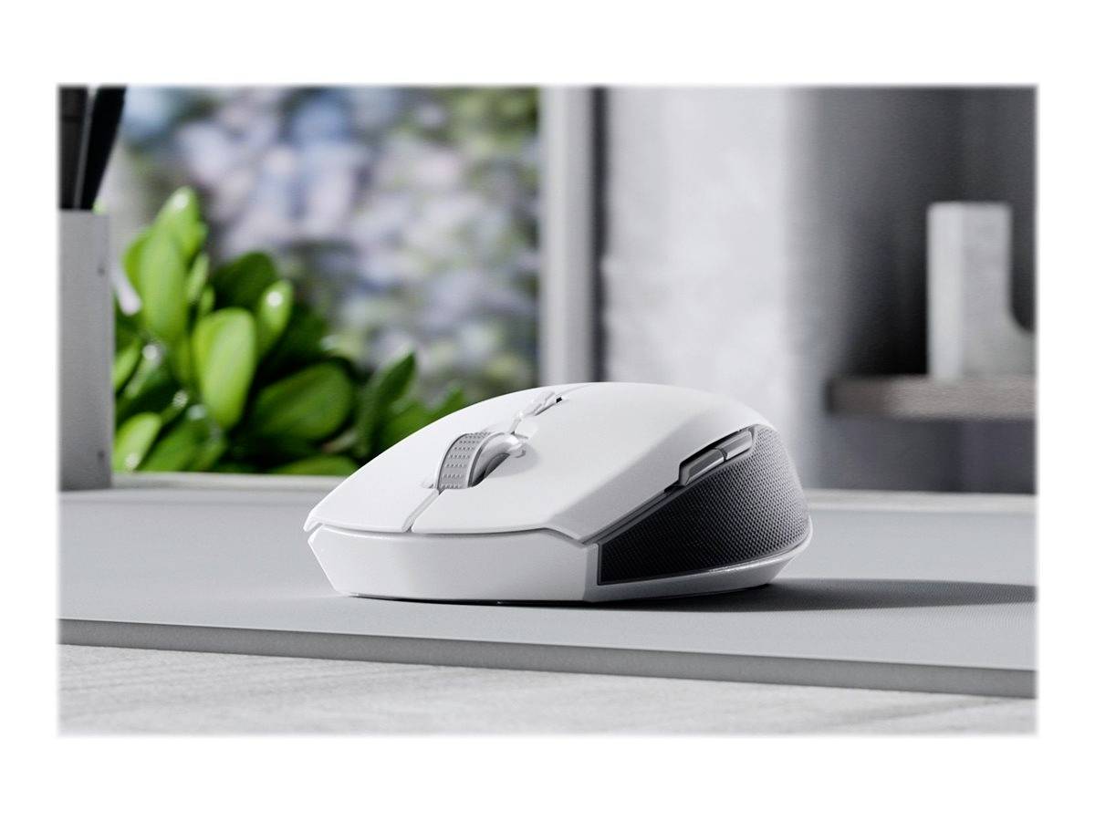 Une souris d'ordinateur blanche sur une table grise, avec des feuilles vertes floues et des étagères en arrière-plan, dans un environnement moderne baigné de lumière.