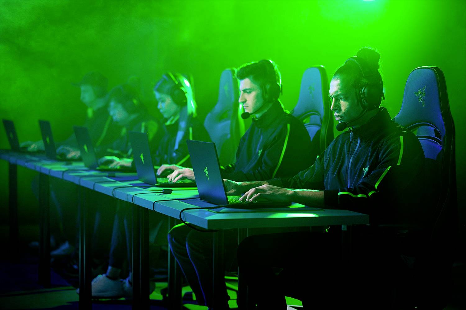 Plusieurs personnes sont assises devant des ordinateurs, portant des casques, et jouent à des jeux vidéo dans une pièce éclairée par une lumière verte.