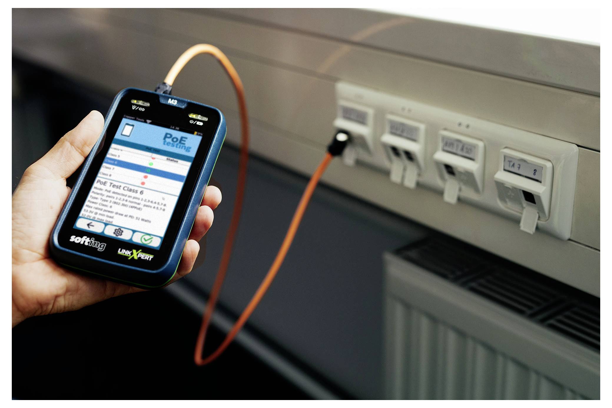 Main qui tient un testeur réseau portatif connecté par un câble orange à des ports Ethernet muraux, l'écran de l'appareil affichant les résultats du test « PoE ».