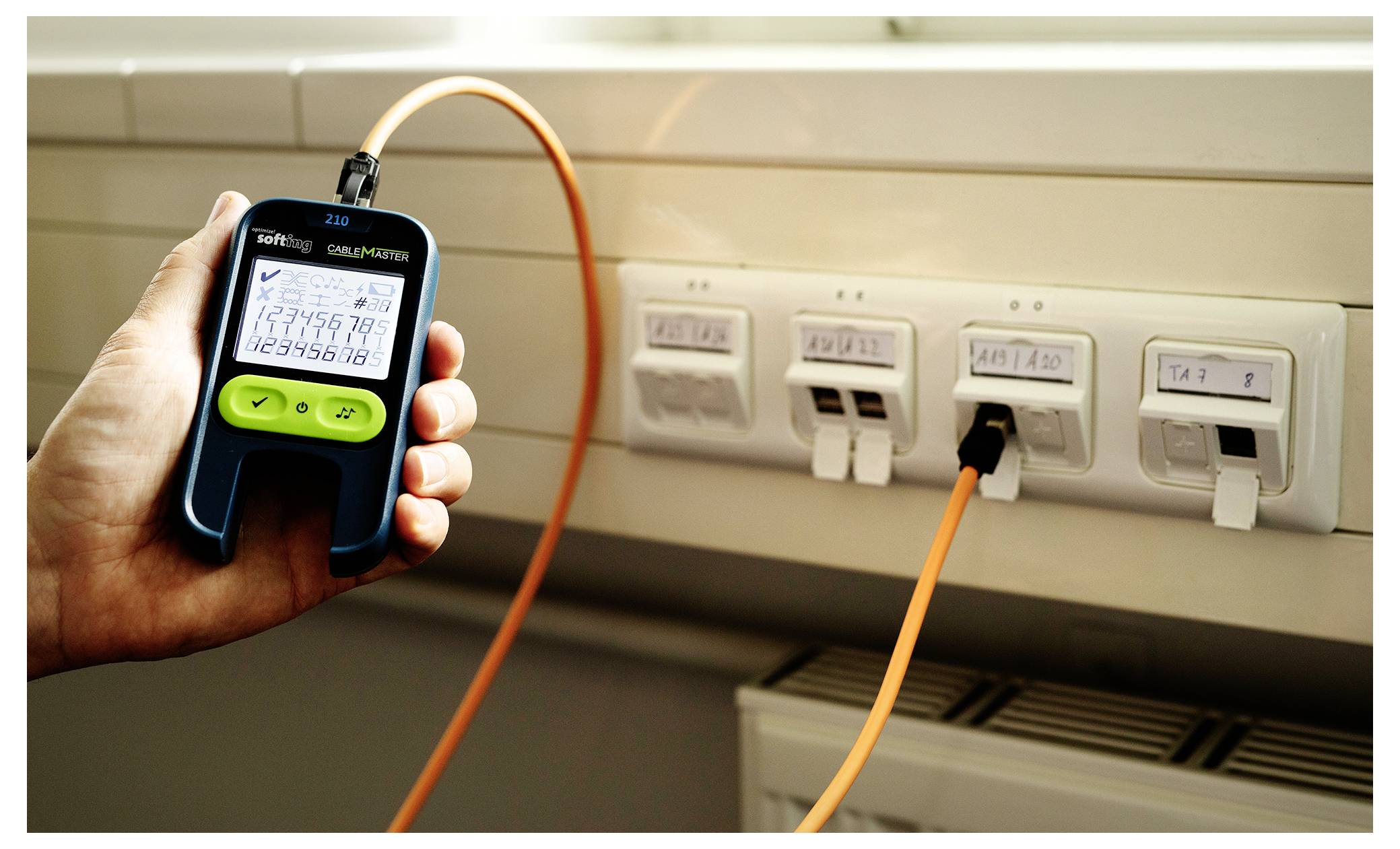 Main tenant une sonde de câble numérique connectée par un câble orange à des prises réseau murales, vérifiant le câblage dans une pièce.