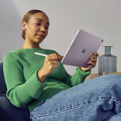 Une personne portant un pull vert est assise sur un canapé et utilise un stylet sur une tablette. Un récipient en verre est visible en arrière-plan.