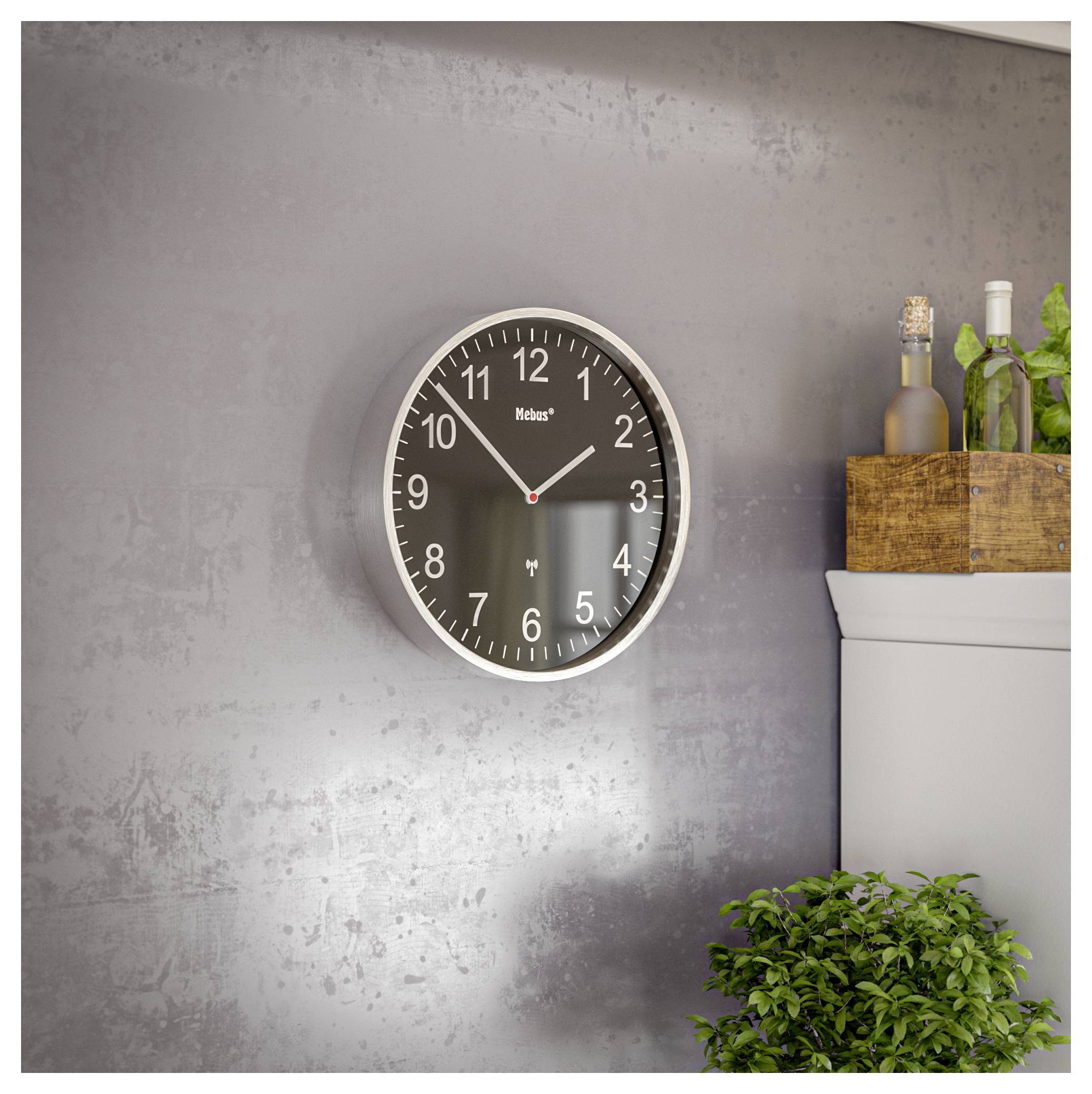 Une horloge murale ronde avec des chiffres blancs et des aiguilles sur un cadran foncé, affichant 10:10. Elle est accrochée sur un mur gris au-dessus d'un meuble avec des bouteilles et de la verdure.