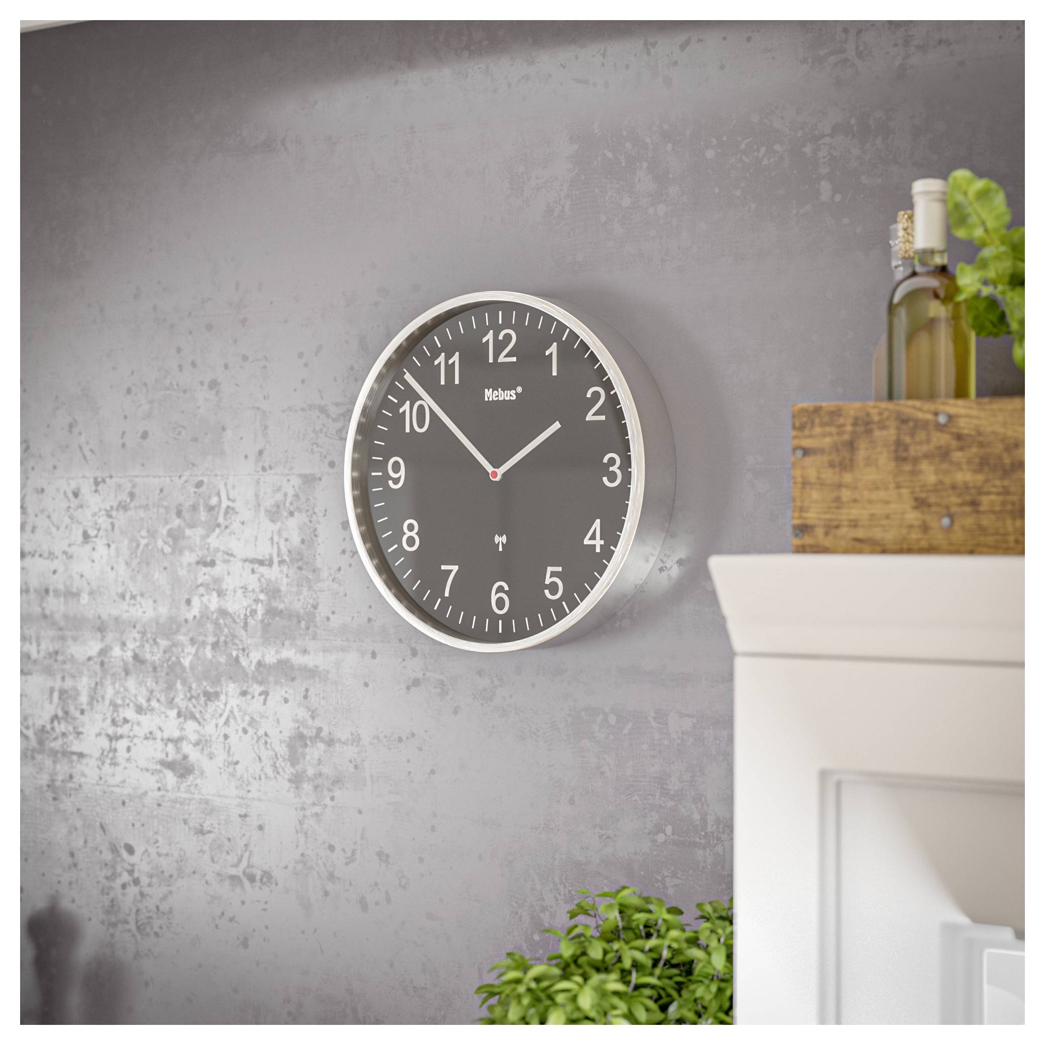 Une horloge murale grise avec des chiffres et des aiguilles blancs est montée sur un mur gris texturé, avec une plante et des bouteilles visibles sur une étagère à proximité.