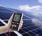 Une main tient un appareil de mesure de panneau solaire devant un grand panneau solaire ; l'appareil affiche des valeurs électriques.