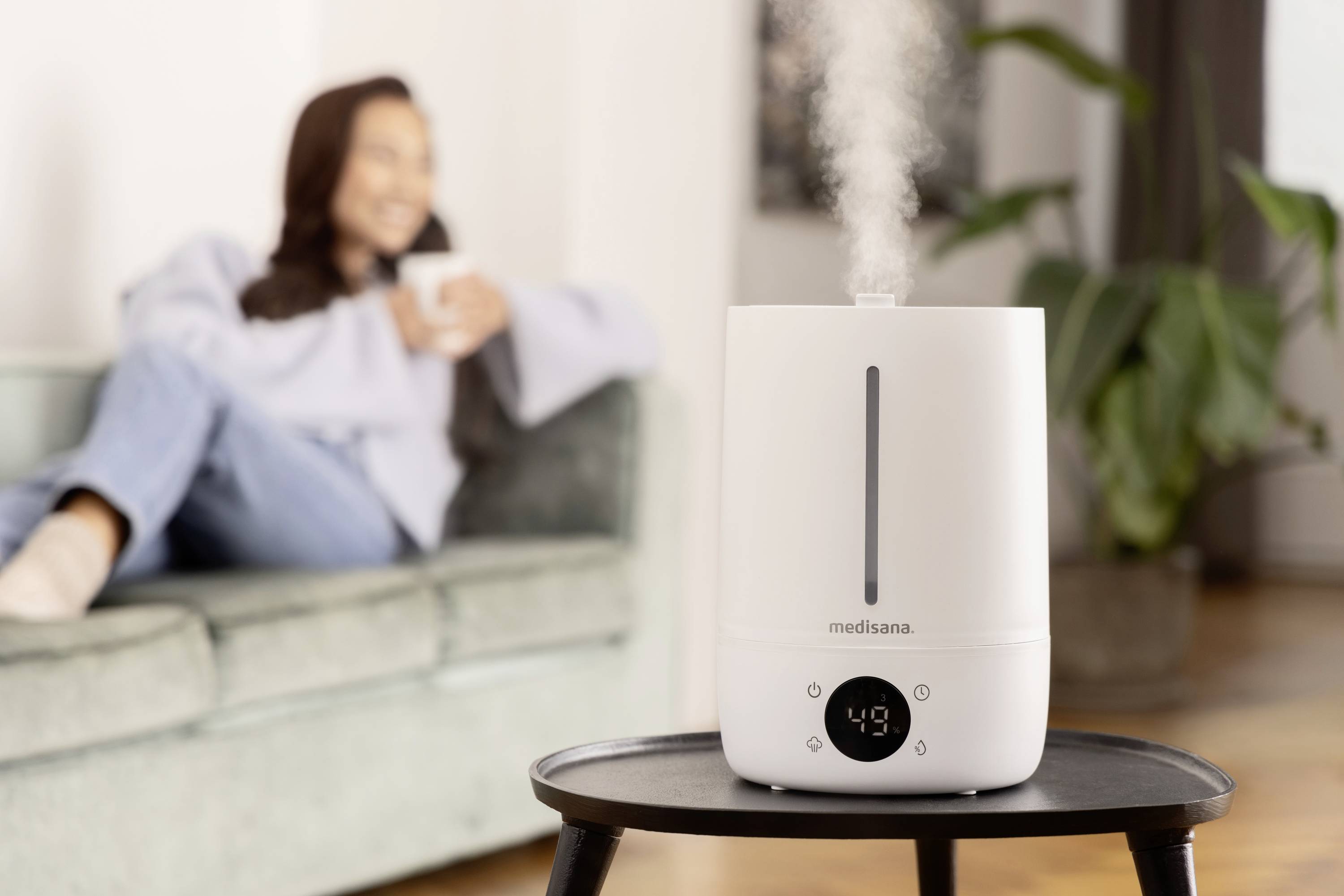 Un humidificateur sur une table émet de la vapeur. En arrière-plan, on aperçoit une personne floue assise sur un canapé.