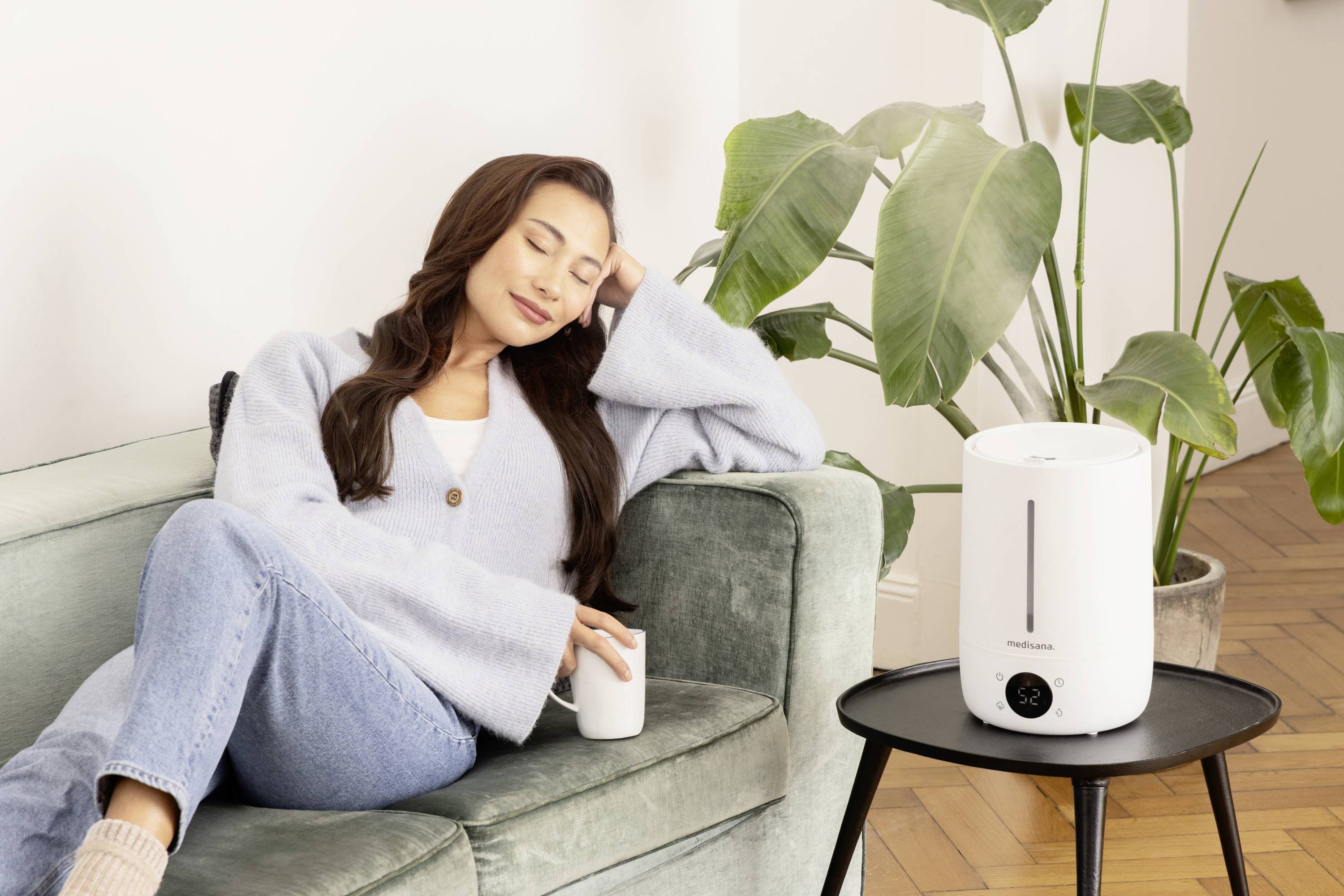 Une femme est assise confortablement sur un canapé avec une tasse, tandis qu'un humidificateur dégage de la vapeur sur une table à côté. Une plante d'intérieur est visible en arrière-plan.