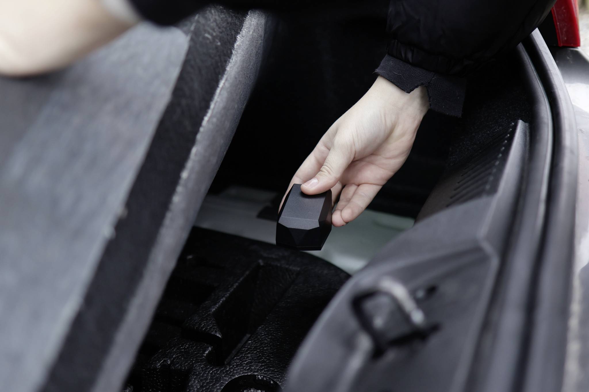 Une main saisit un objet noir à l'intérieur du coffre d'une voiture. La scène montre le rangement ou le retrait d'un objet de l'espace de stockage.