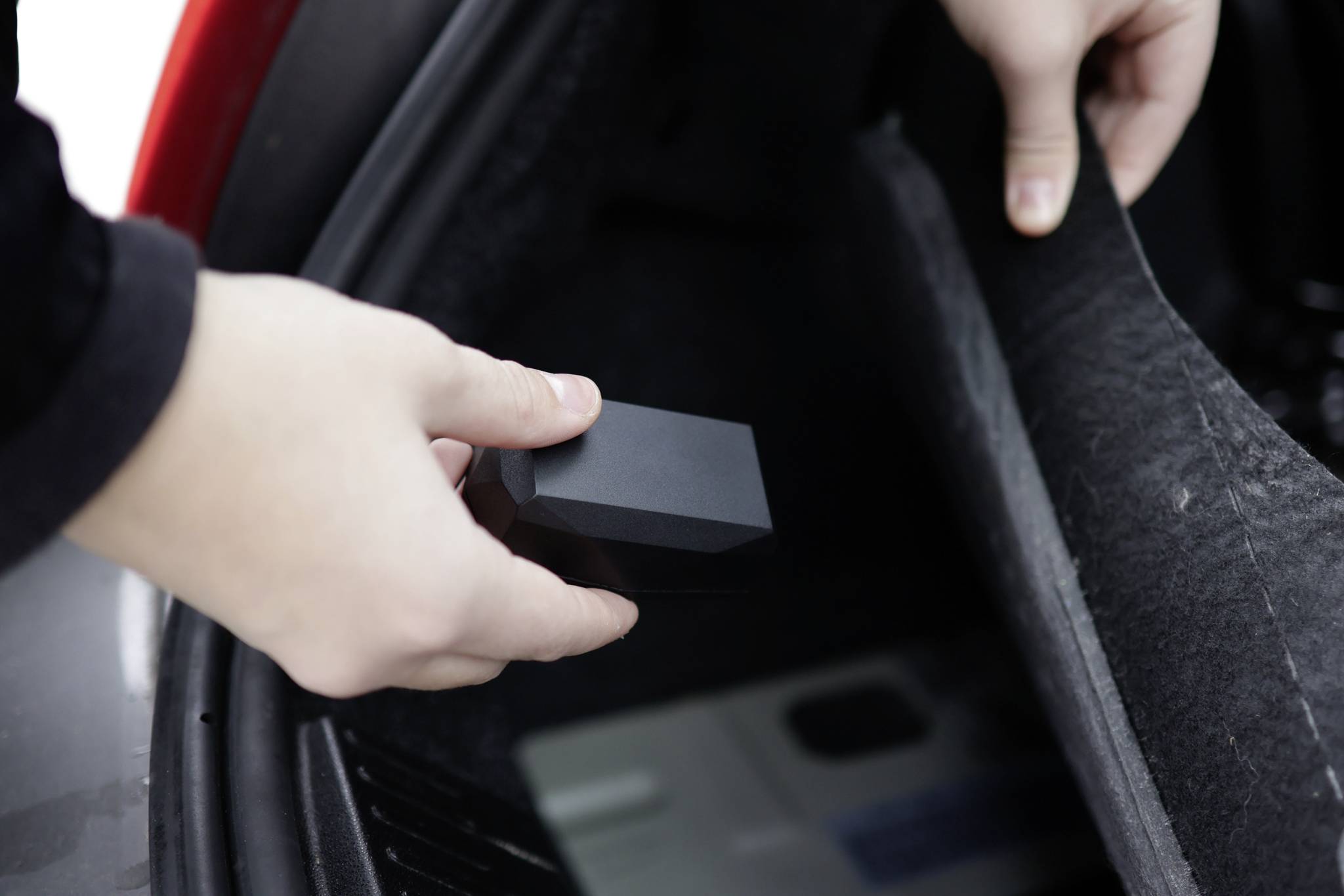 Une personne place un petit appareil noir dans le coffre d'une voiture. L'appareil pourrait être un GPS tracker ou un dispositif électronique similaire.