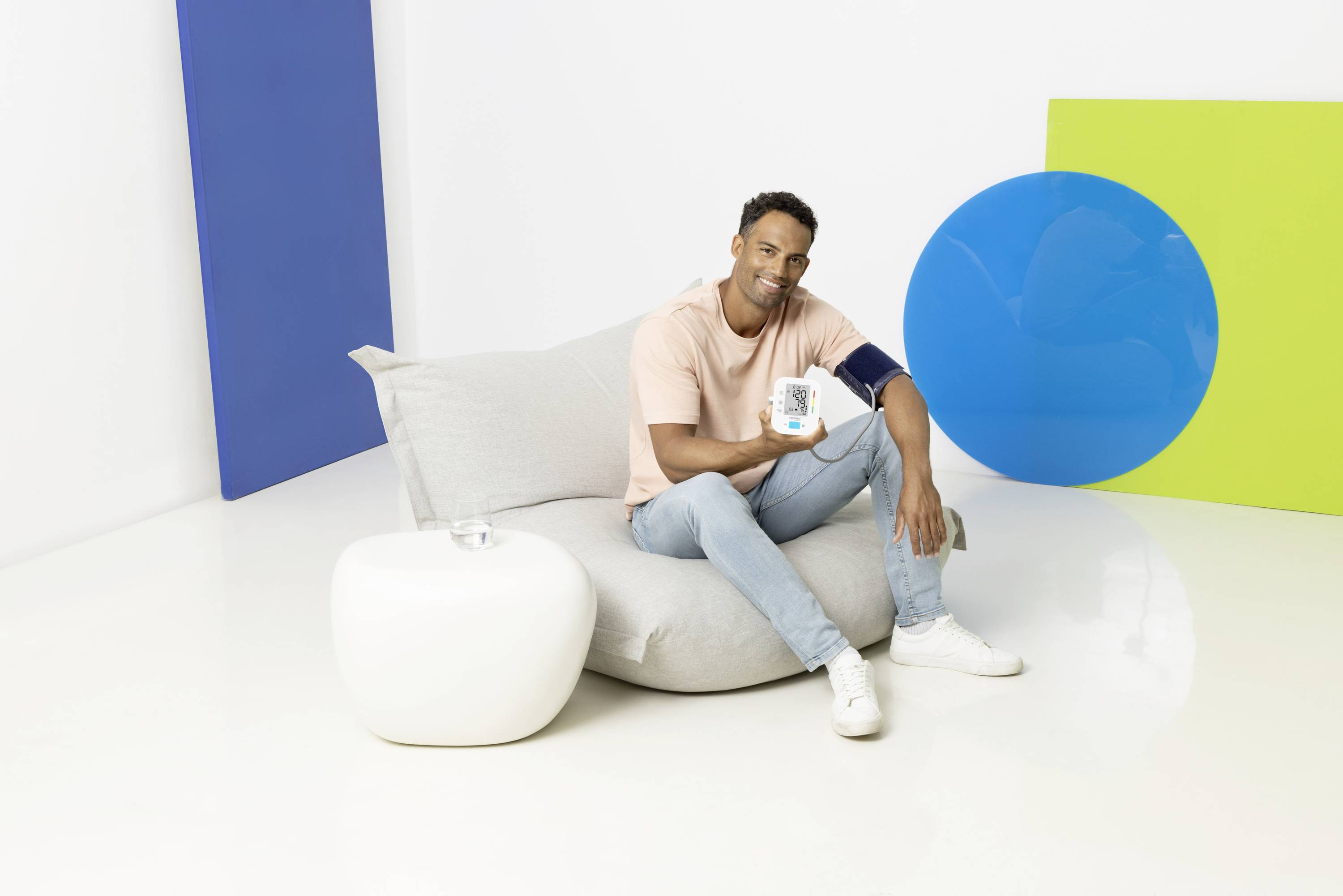 Une personne est assise sur un fauteuil gris et tient un appareil de mesure de la tension artérielle. À l'arrière-plan, des formes bleues et vertes sont disposées sur un mur blanc.