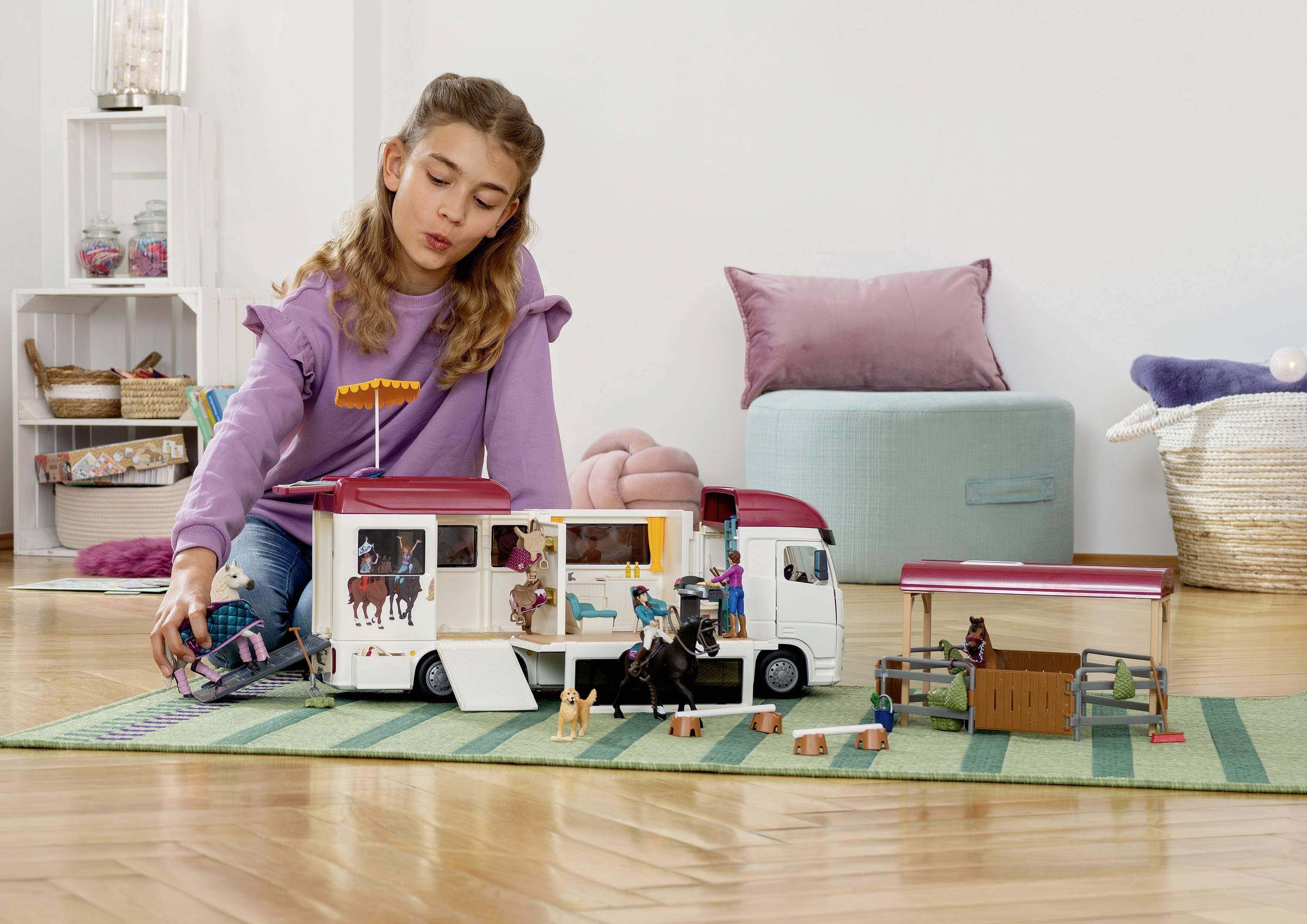 Une petite fille joue sur un tapis vert avec un camion-transport de chevaux et des écuries de jouets. En arrière-plan, on peut voir des coussins et un panier tressé.