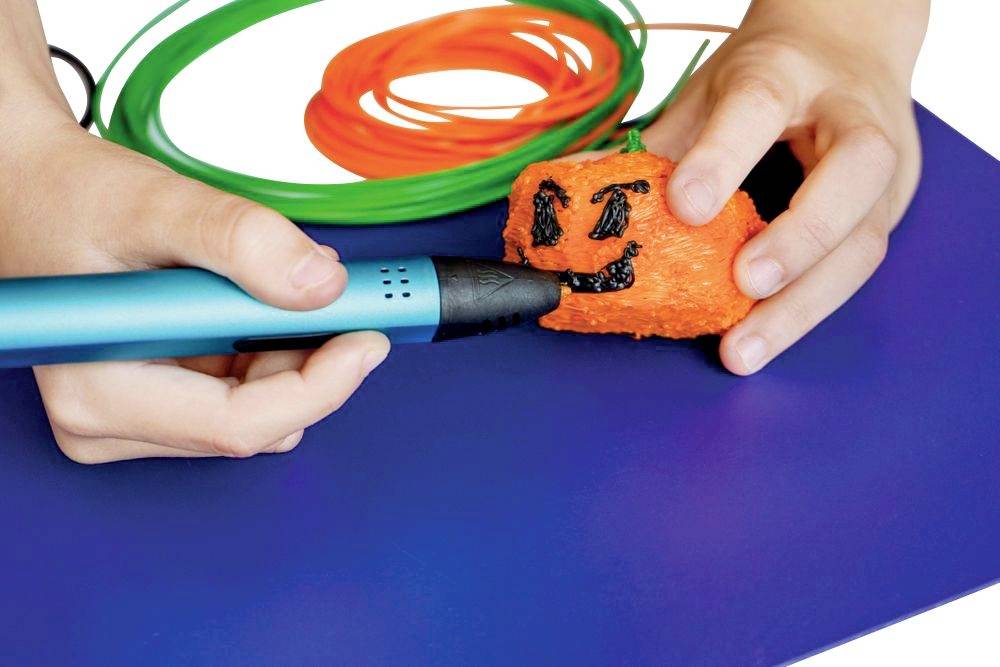 Une main dessine un visage sur une petite citrouille orange à l'aide d'un stylo 3D. Des fils colorés sont disposés en arrière-plan.